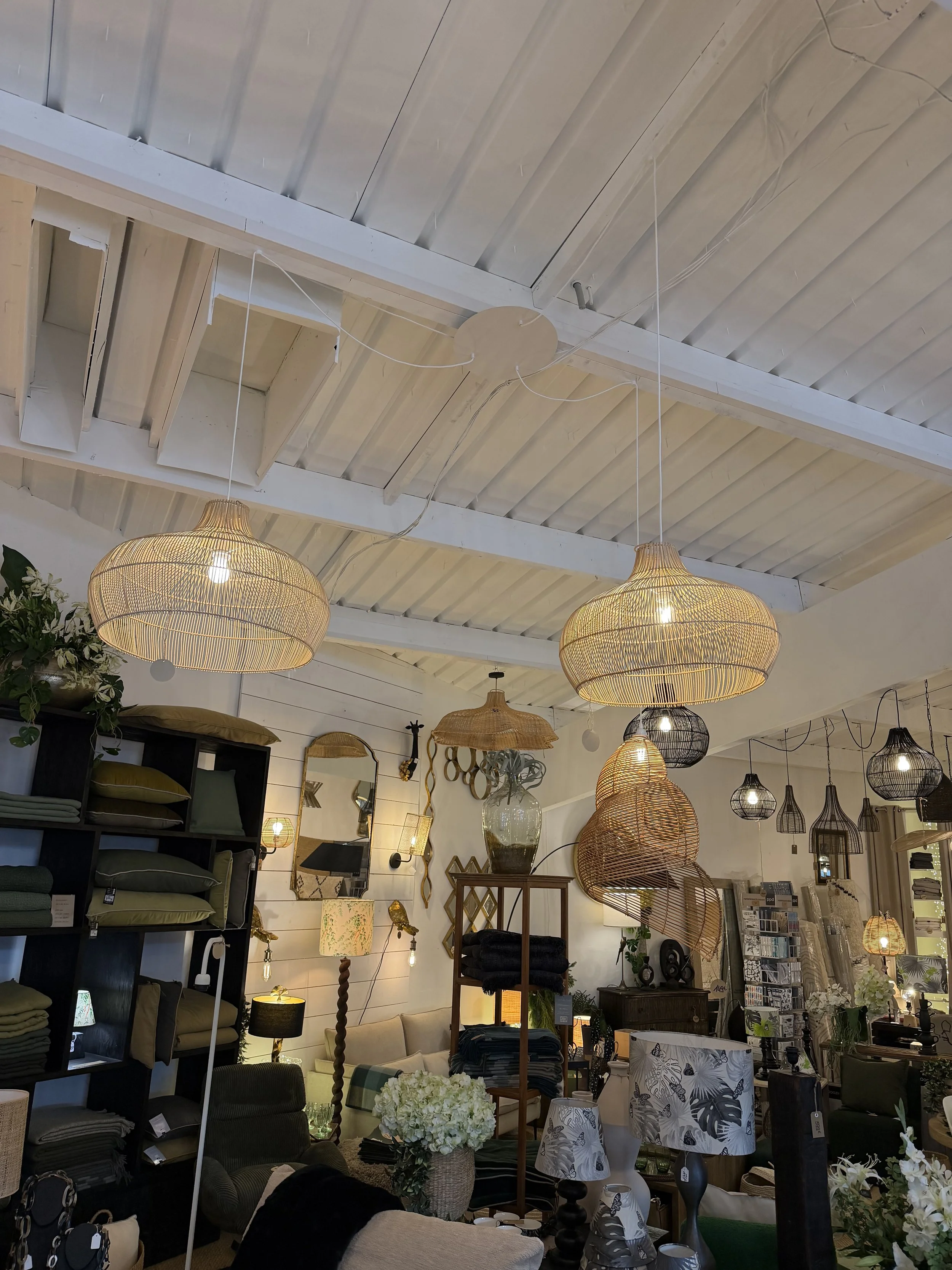 L'intérieur d'une boutique decorée avec des luminaires en rotin suspendus, des étagères avec des coussins, des lampes et des accessoires décoratifs.