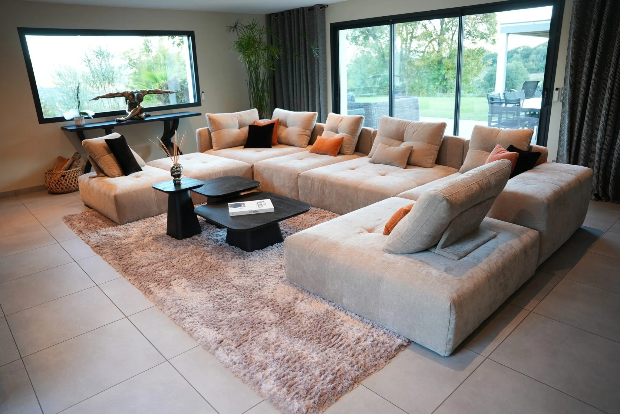 Salon moderne avec canapé beige en forme de U, tapis beige, deux tables basses noire, décorations avec coussins noirs et orange, grande fenêtre avec vue sur jardin, porte-fenêtre avec terrasse extérieure.