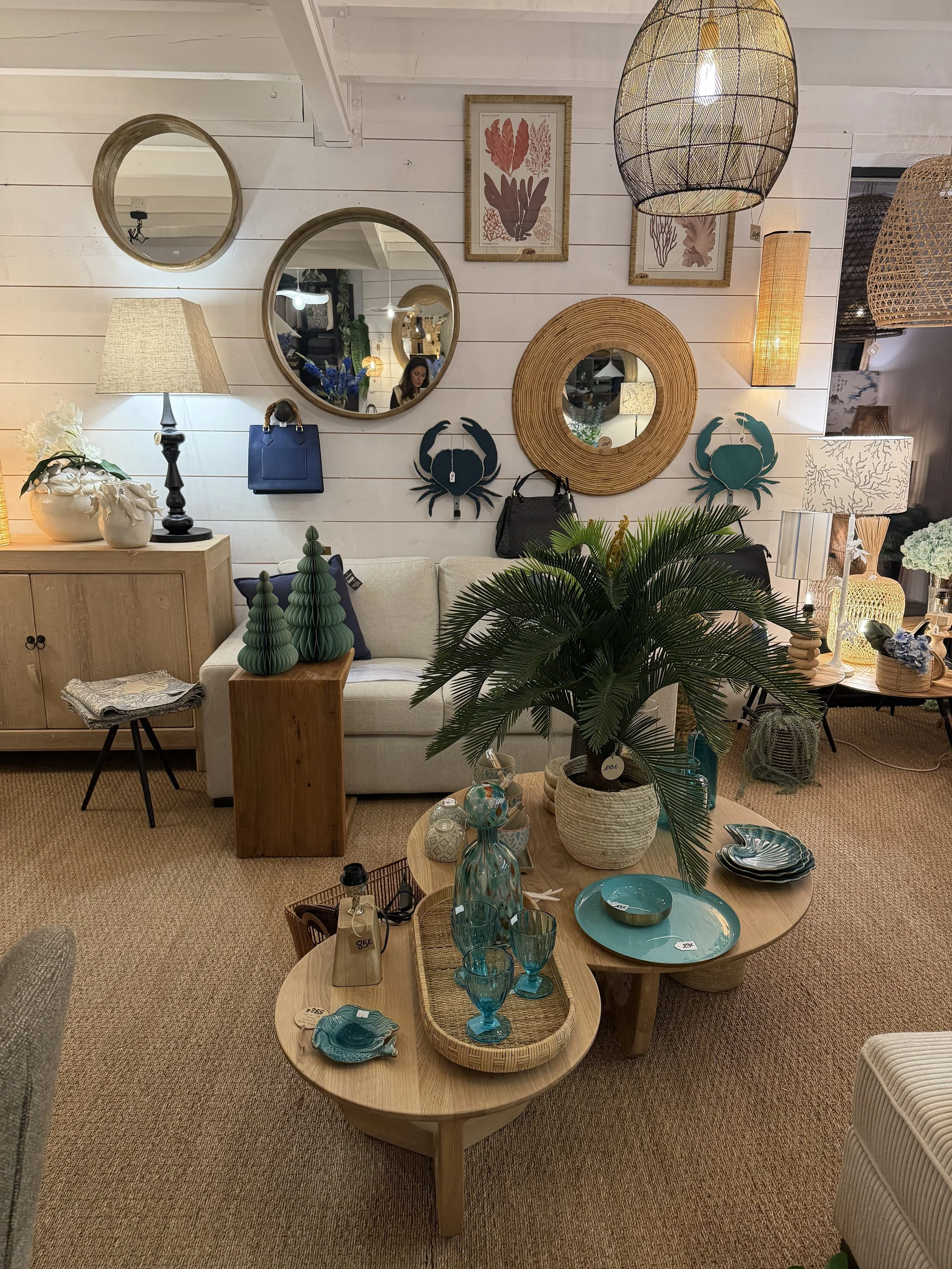 Une décoration intérieure avec un mur blanc en bois, tableaux, miroirs, lampes, des plantes, et un mobilier en bois avec des objets décoratifs, notamment des vases en verre bleu, assiettes en turquoise, et des accessoires divers.