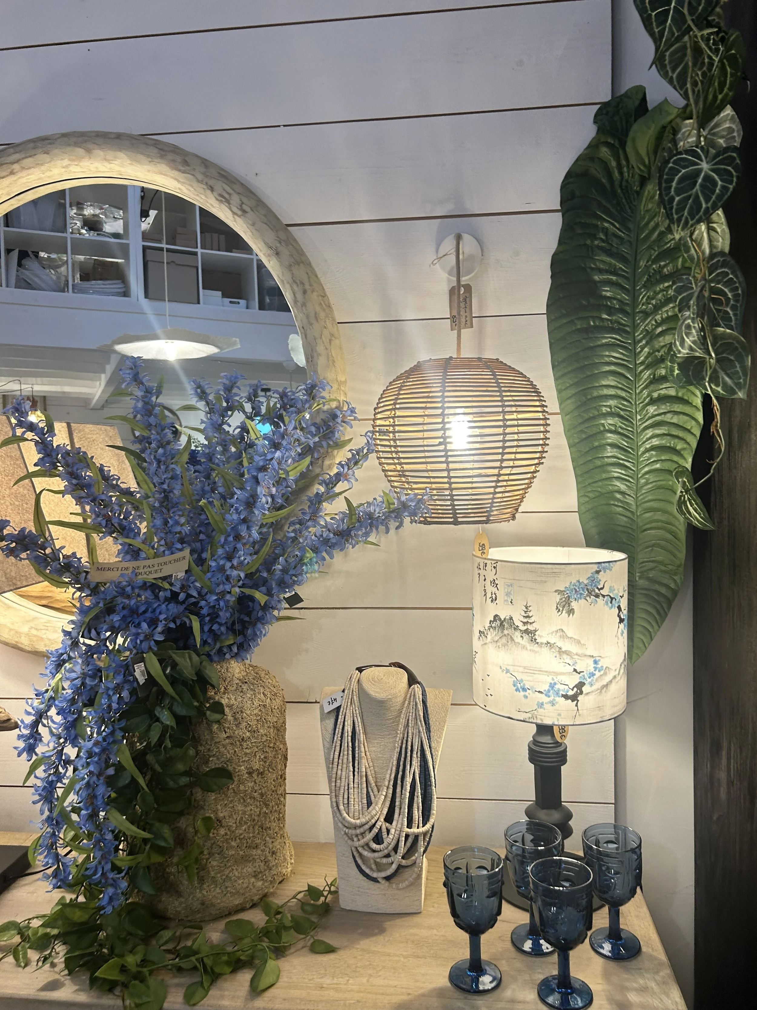 Une décoration intérieure comprenant un miroir rond, un vase avec des fleurs violettes, une lampe à abat-jour japonisante, un collier de perles sur un support, une lampe avec un motif décoratif, des verres à vin bleus, et de grandes feuilles vertes
