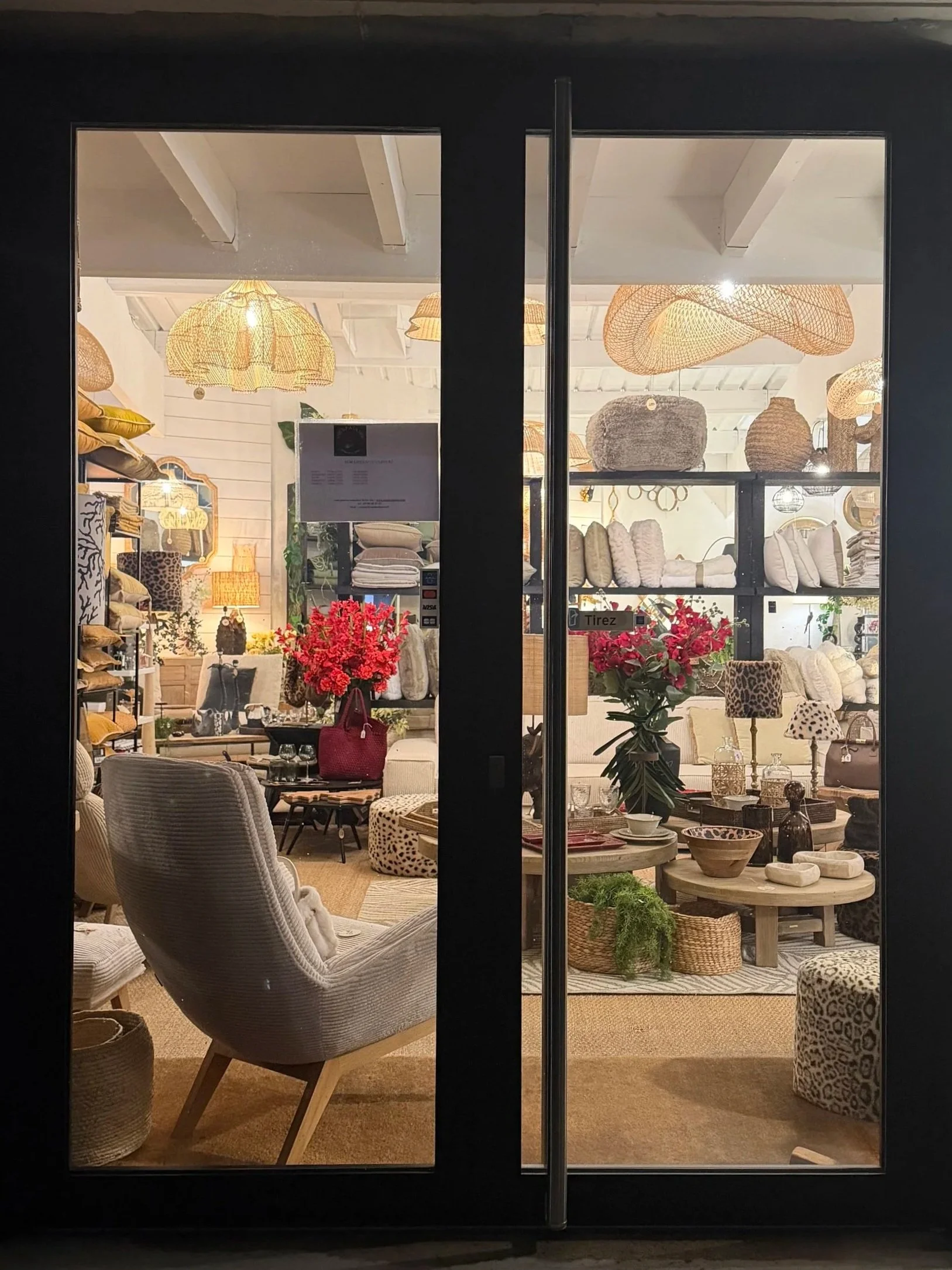 Une vitrine d'un magasin de décoration intérieure avec un intérieur éclairé, comprenant des lampes en rotin, des coussins, des tables en bois, des plantes, et des objets décoratifs variés.
