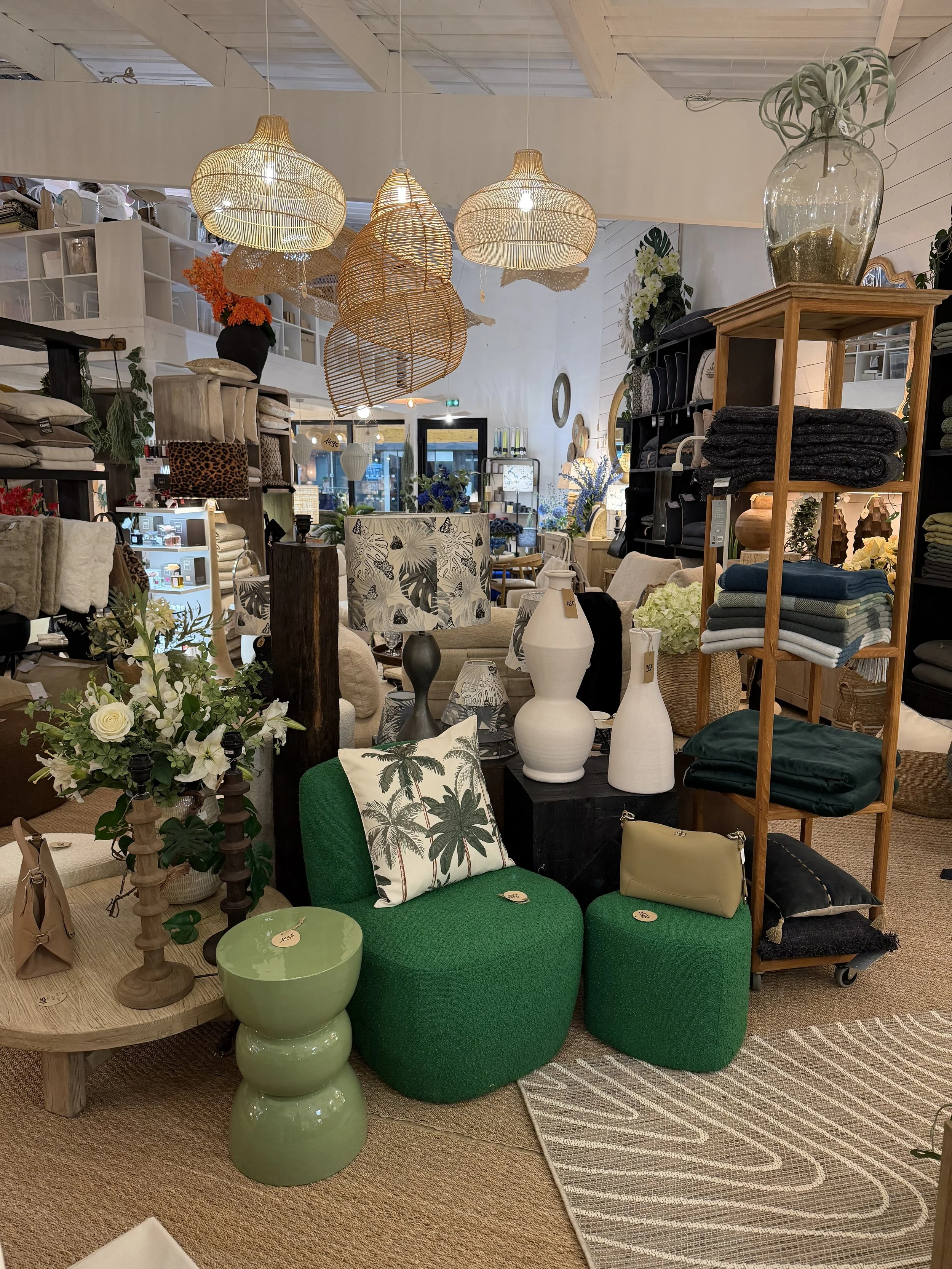 Intérieur d'un magasin de décoration avec des meubles, des luminaires en matériau naturel, des vases blancs, un fauteuil vert avec un coussin à motifs de palmiers, et diverses hampes avec des plantes et des textiles.