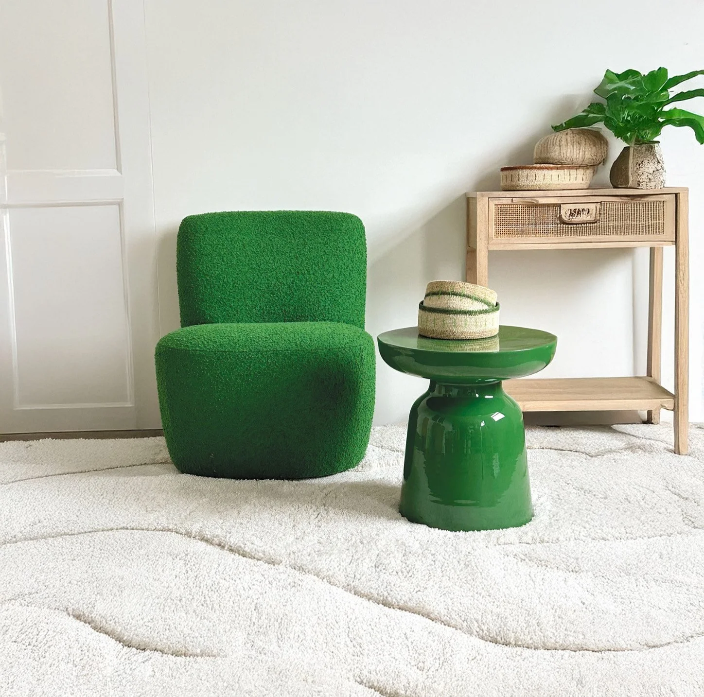 Un fauteuil vert en tissu, une table verte en céramique, une console en bois avec des paniers et une plante verte