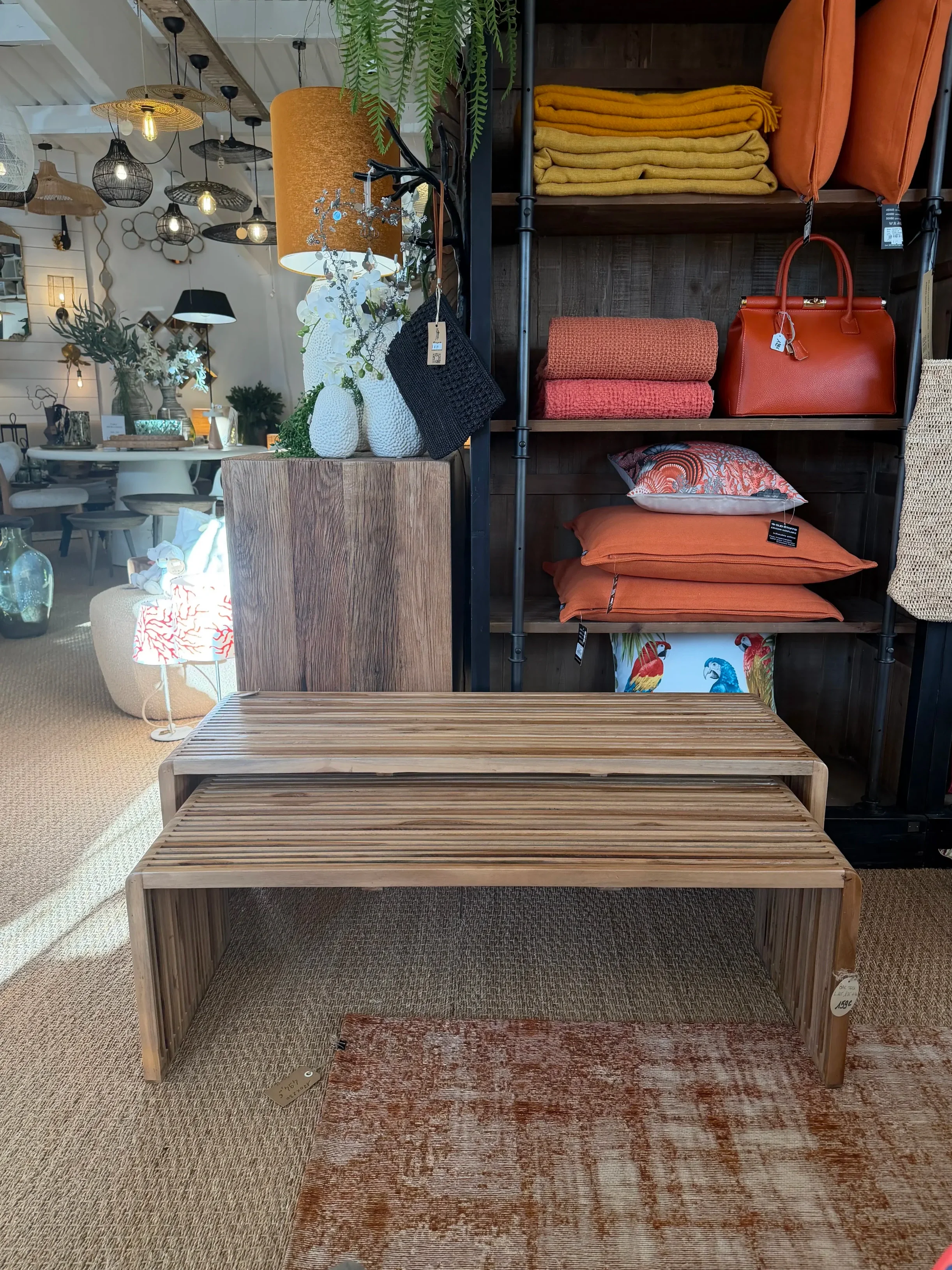 Deux bancs en bois clair dans une boutique décorative, avec étagères contenant des coussins, des sacs à main et des couvertures colorées, derrière un plateau en bois et un lampadaire orange.