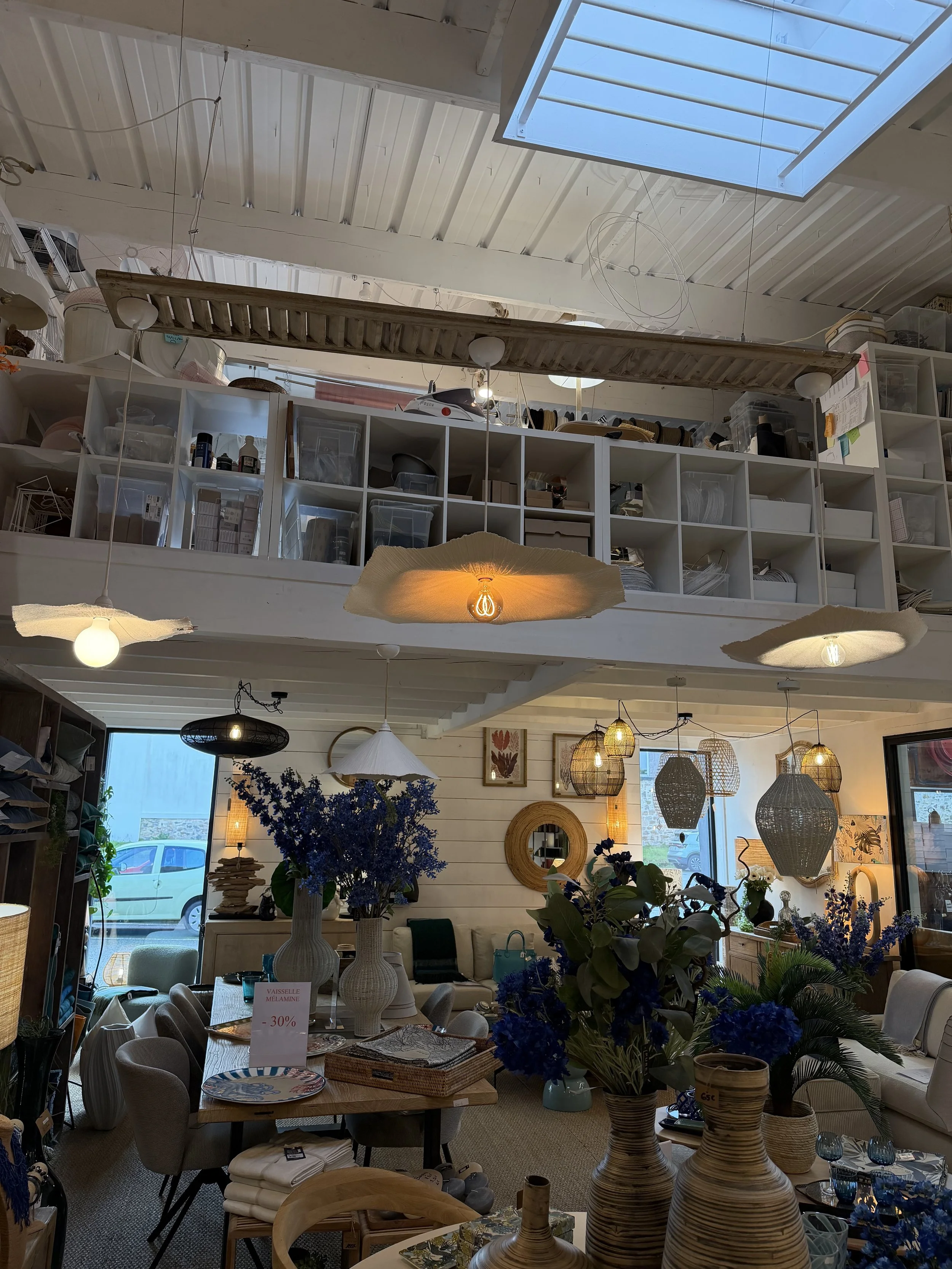 Intérieur d'un magasin ou showroom avec mobilier et décoration, notamment des vases avec des fleurs bleues, un tableau avec affichage promotionnel, luminaires suspendus et une disposition de canapés et d'objets décoratifs.