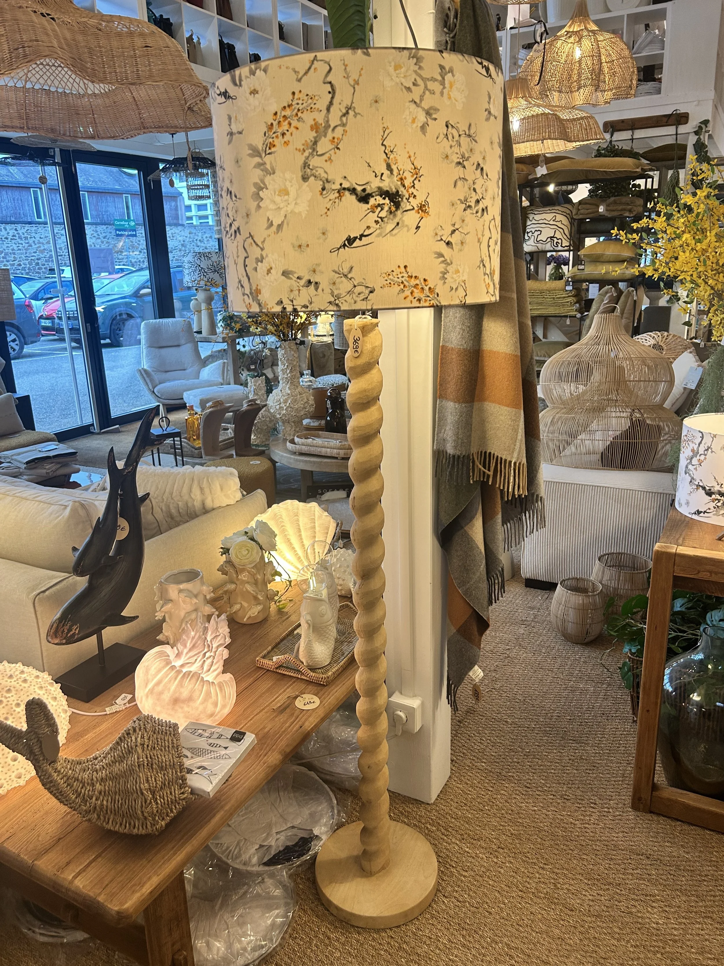 Lampe sur pied en bois avec un abat-jour motif floral dans un magasin de décoration, entourée d'objets décoratifs.