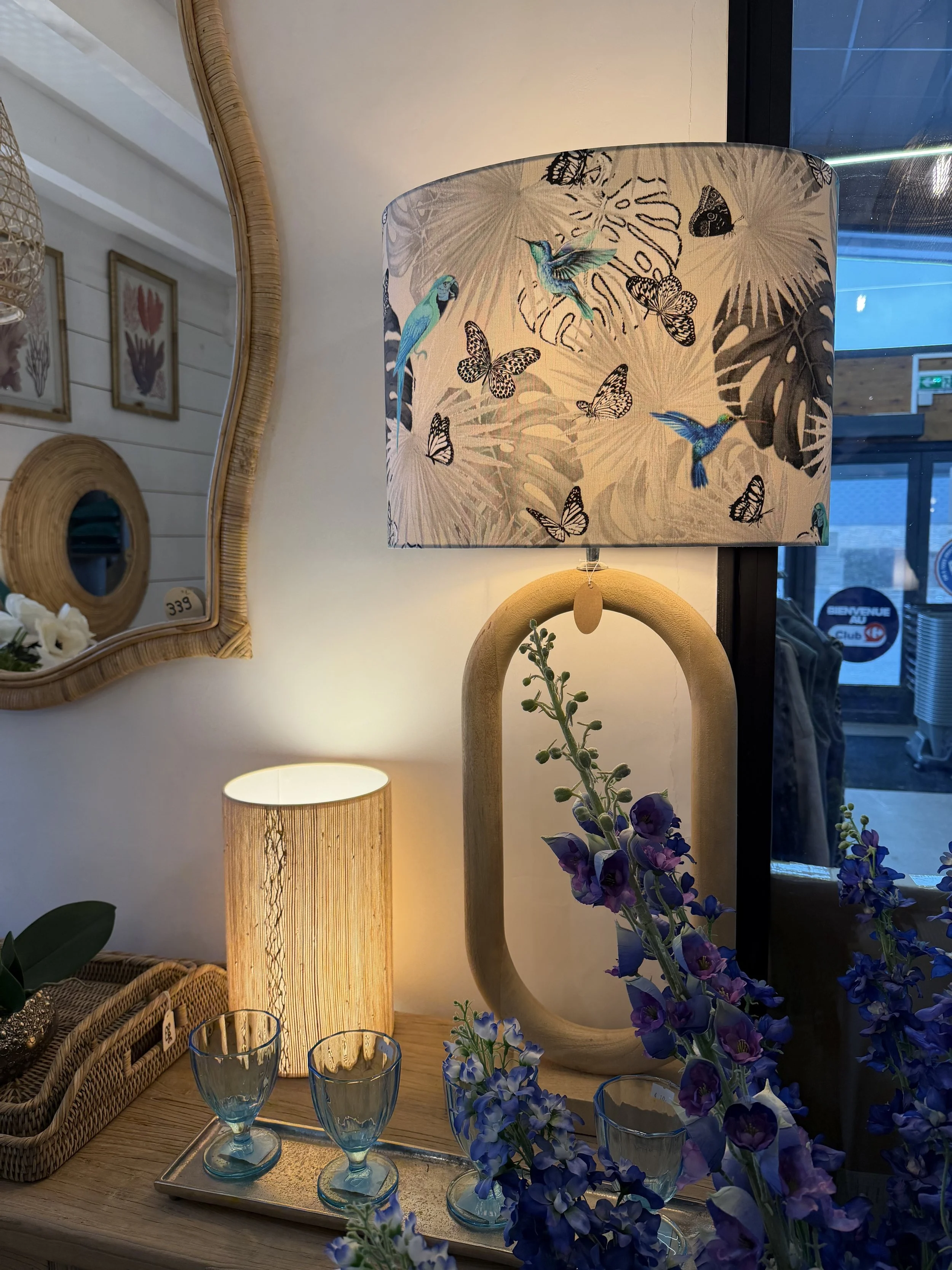 Deux lampes décoratives, une avec un abat-jour à motifs de colibris et papillons, l'autre allumée, sur une table avec des verres en dessous. Des fleurs violettes et un miroir sont aussi visibles.