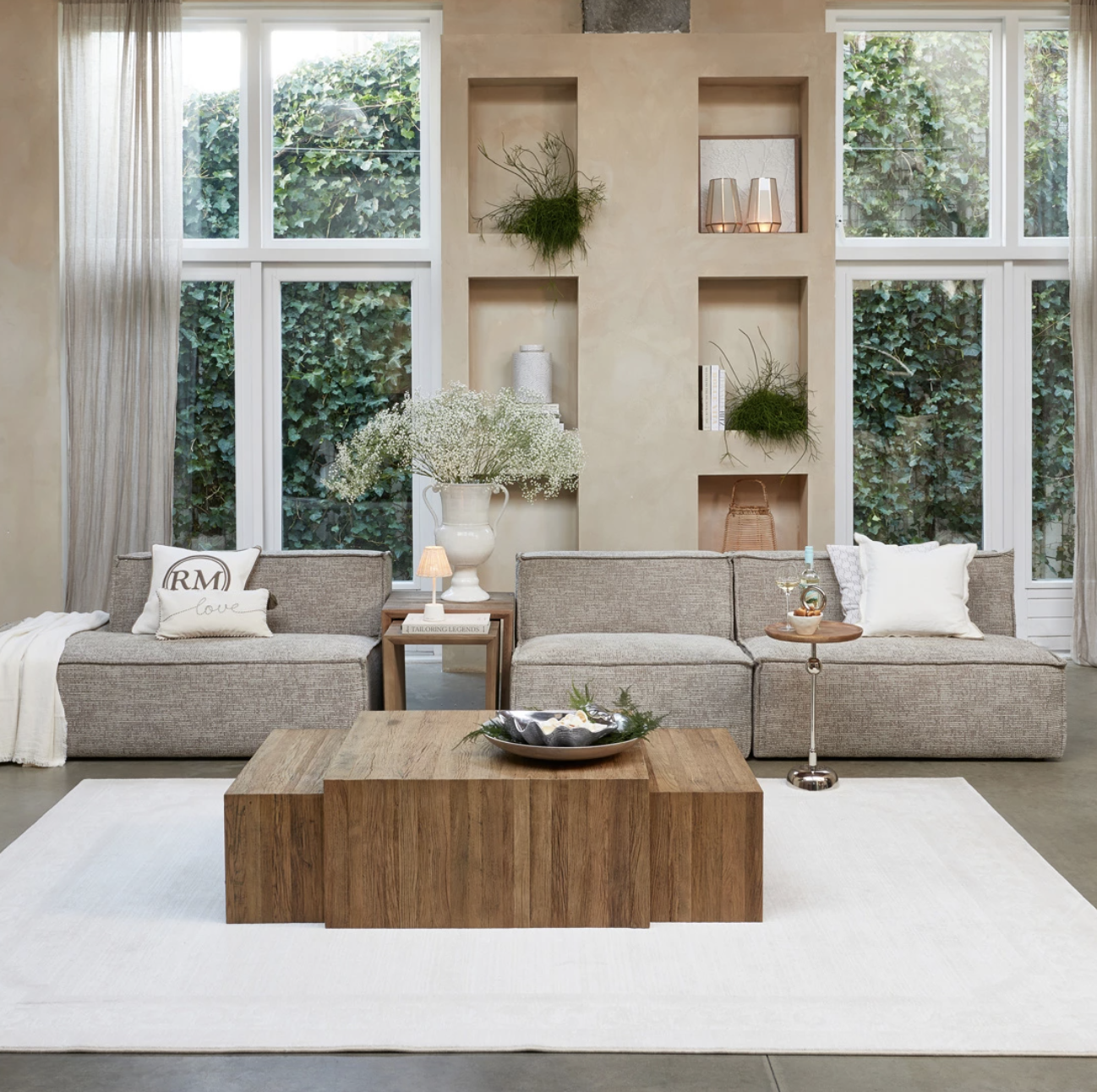 Salon moderne avec deux canapés gris, une table en bois, un vase de fleurs blanches, des décorations murales, et de grands fenêtres avec vue sur une haie verte.