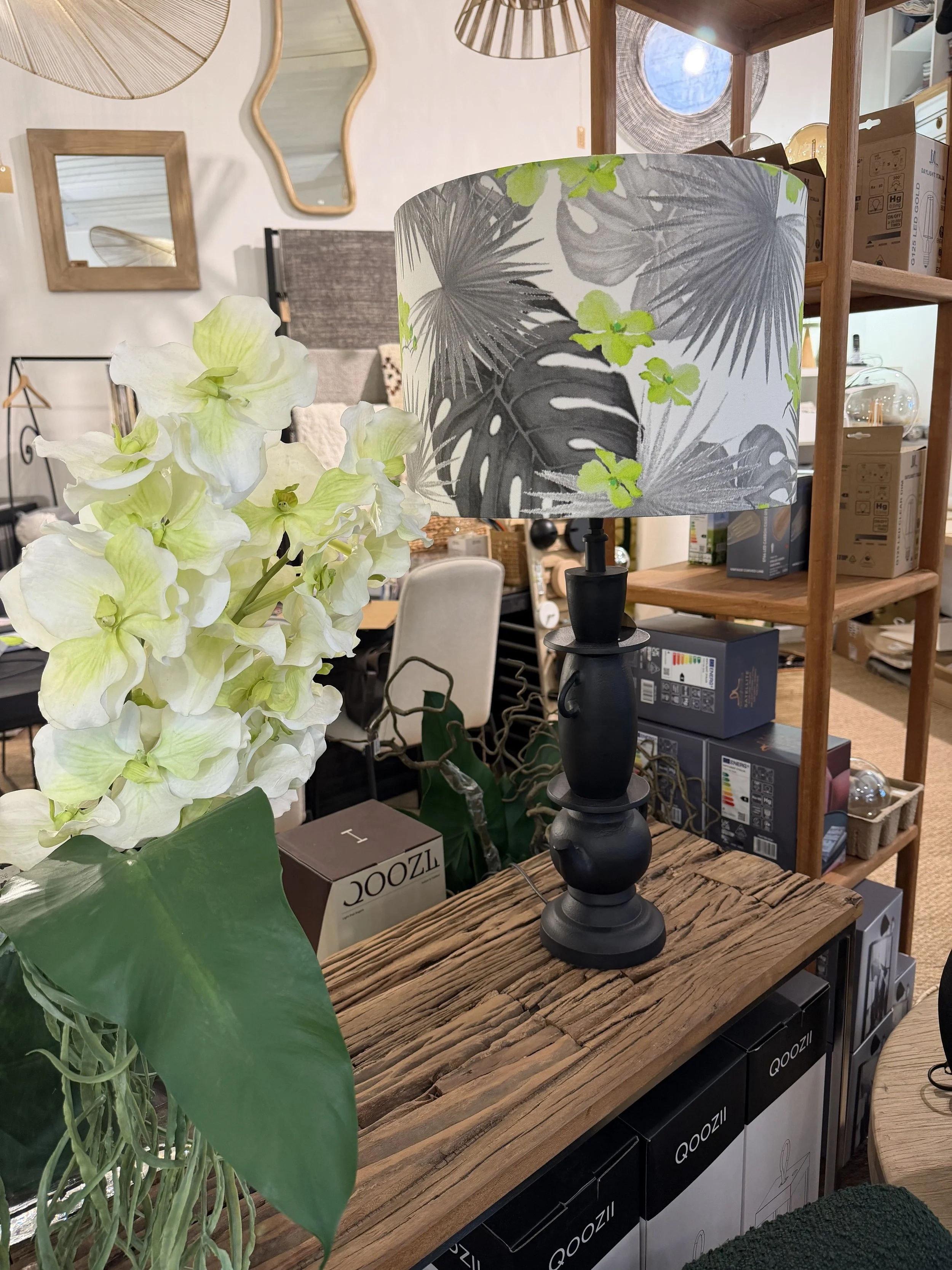 Lampe de table noire avec un abat-jour à motif de feuilles tropicales verts et gris, placée sur une table en bois rustique dans un magasin de décoration. Autour, des plantes artificielles ou décoratives et des rayons avec des boîtes et objets décorat
