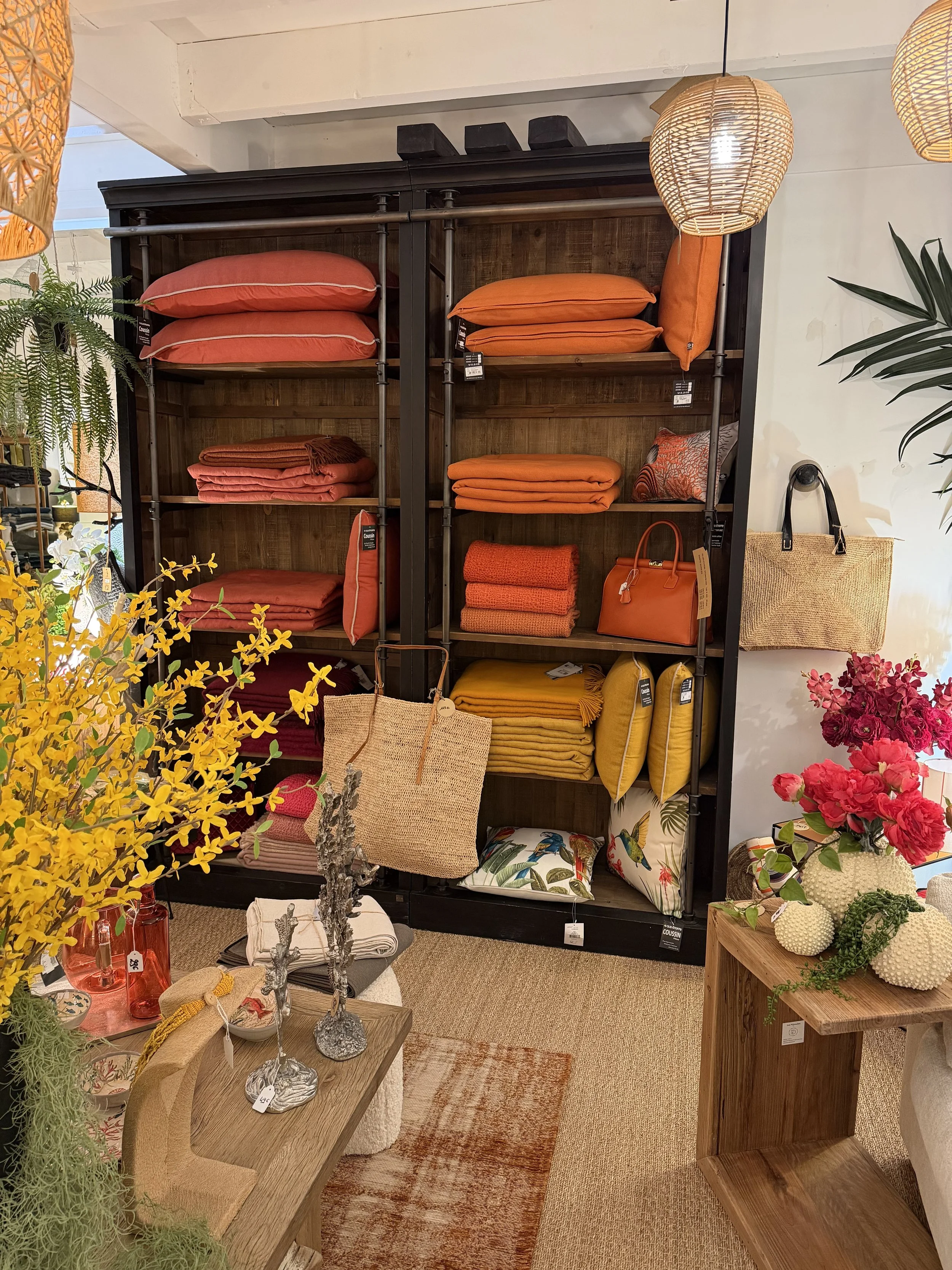 Rayonnage avec des coussins et des plaids orange, rouges et jaunes dans un magasin de décoration intérieure, avec des lampes en papier, des fleurs et des accessoires autour.