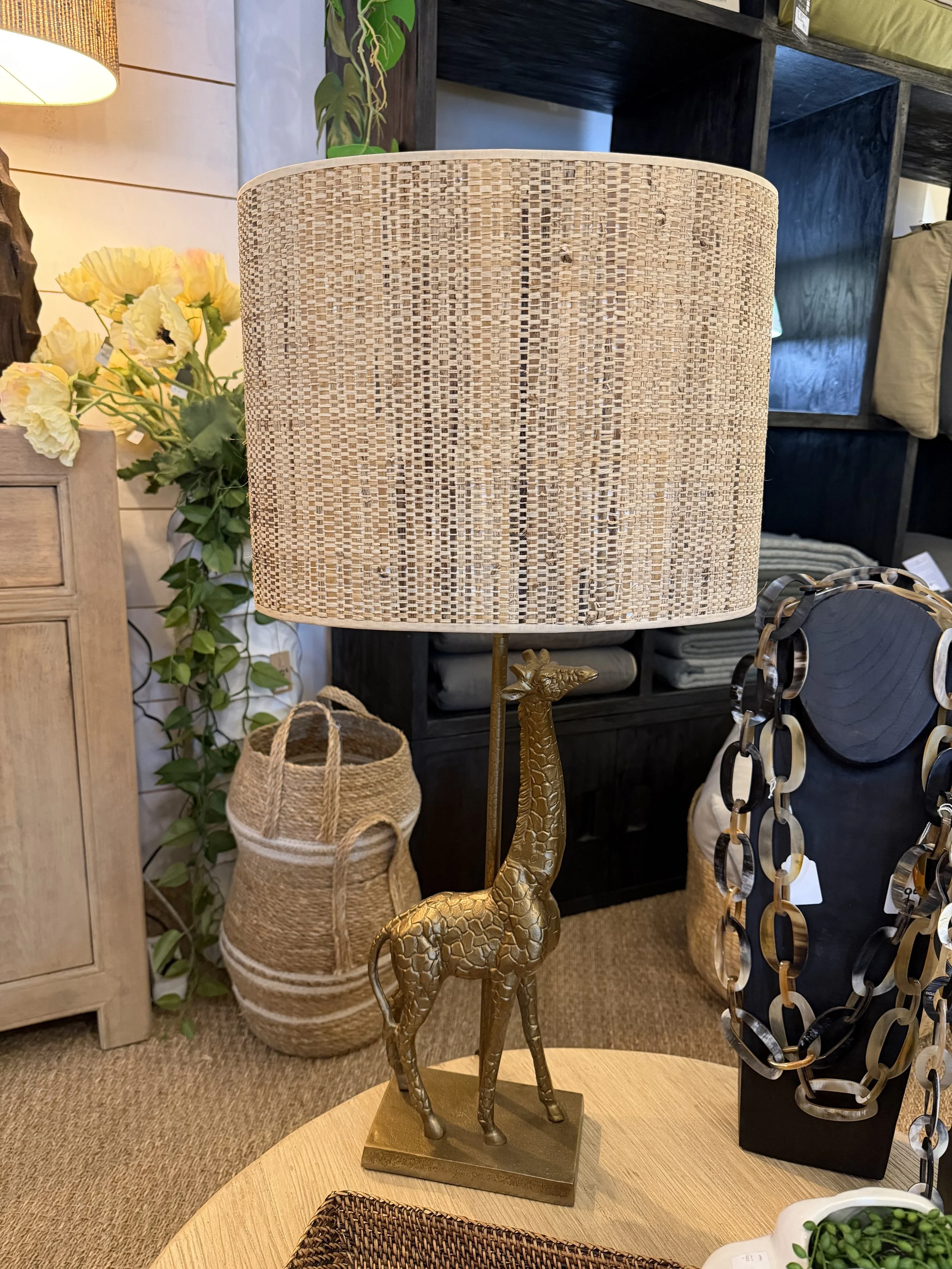 Lampe de table avec un abat-jour en rotin et un pied représentant une girafe dorée, placée sur une table en bois dans une décoration intérieure avec des plantes et des objets décoratifs.