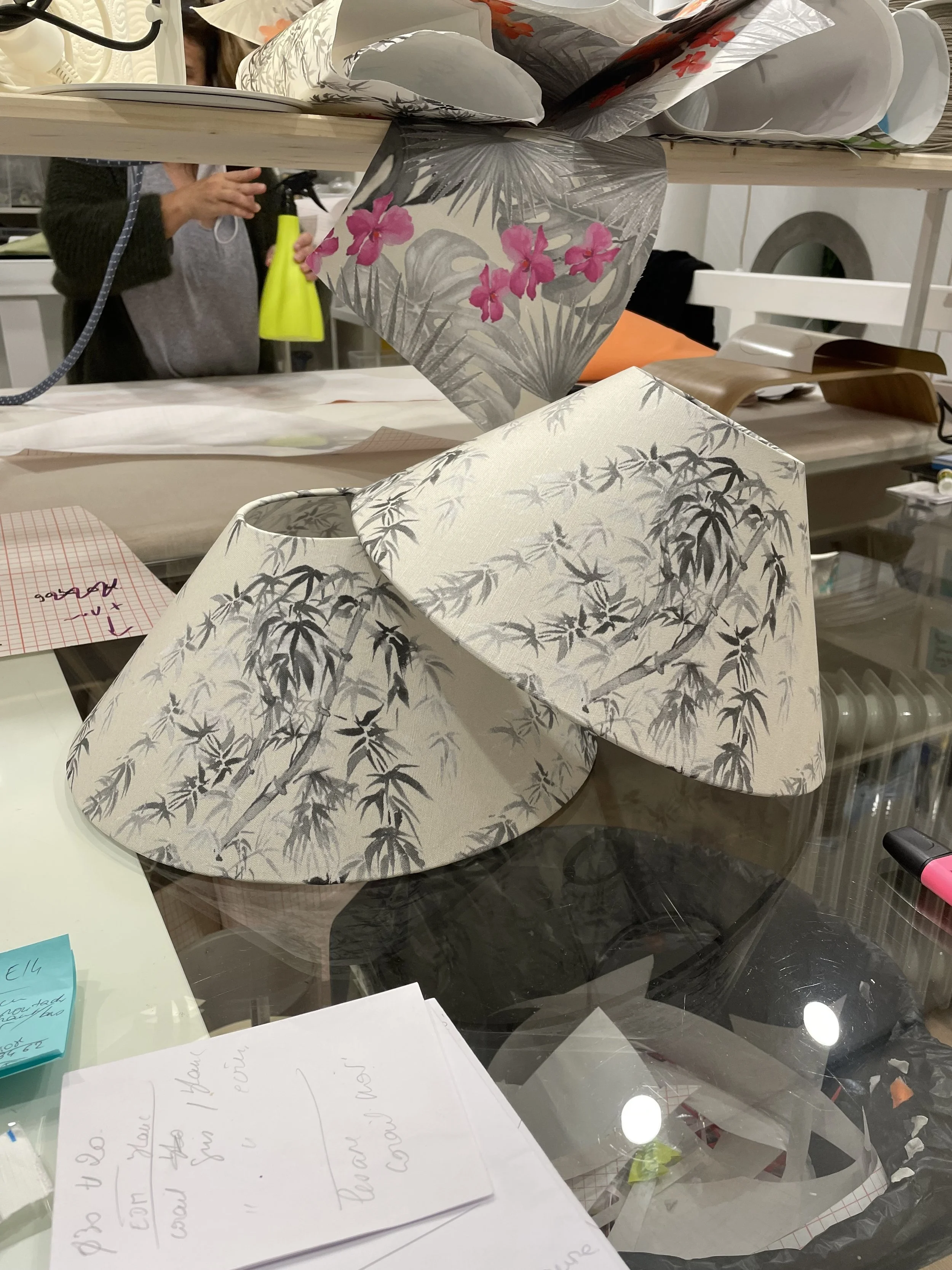 Deux lampes à abat-jour avec motifs de bambous, l'une en noir et blanc, l'autre avec des fleurs roses. Ils sont placés sur une table de travail. En arrière-plan, une personne tient un vaporisateur jaune et il y a divers objets et papiers.