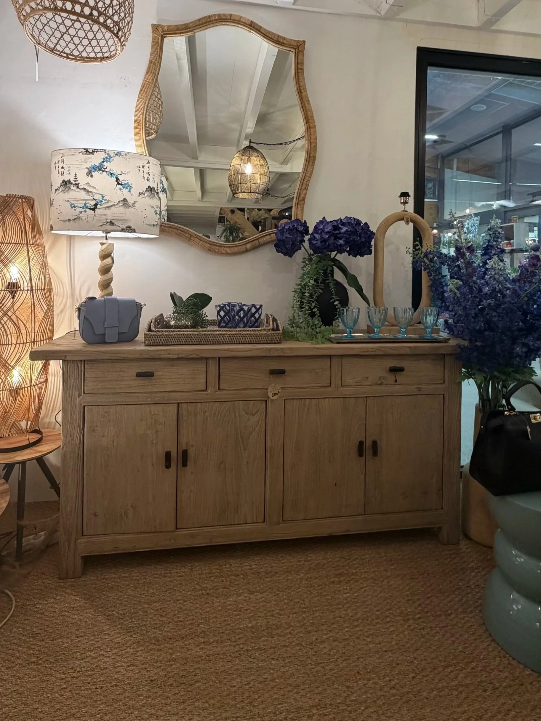 Une commode en bois naturel avec des tiroirs et des portes, décorée de vases avec des fleurs violettes et d'une petite étagère, avec un grand miroir mural en bois au-dessus.