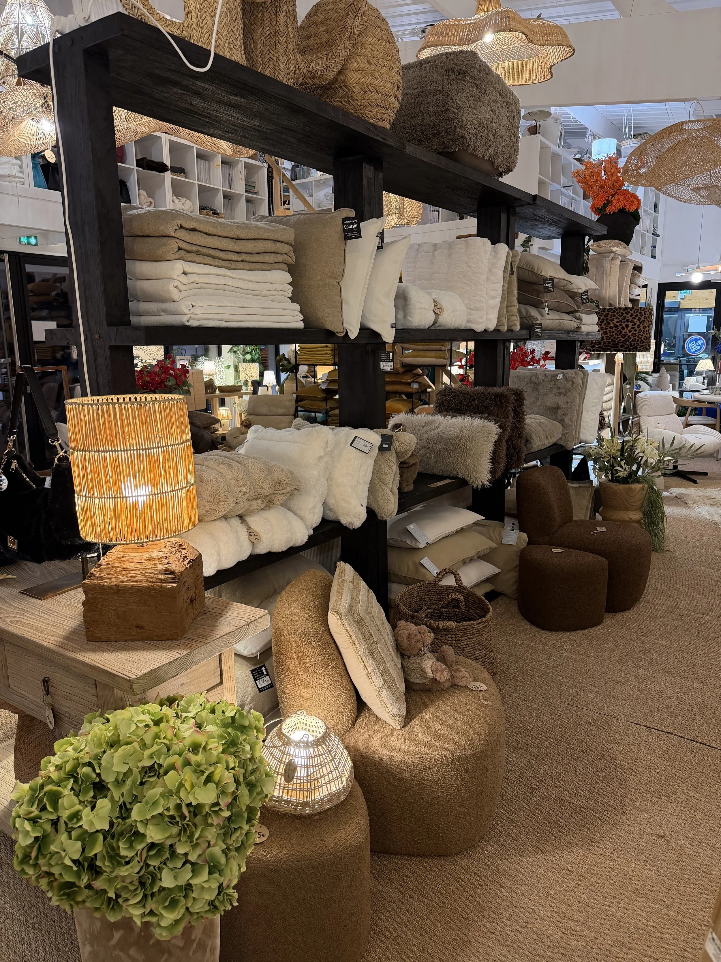 Étagère en bois noir avec des coussins, plaids et décorations en textiles de couleur beige, blanc, marron, et crème dans un magasin de décoration intérieure.