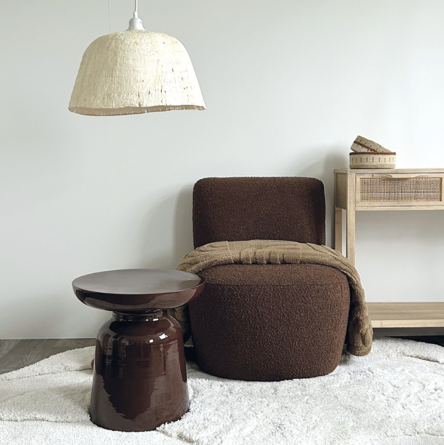 Un coin de salon avec une chaise marron, une petite table en céramique marron foncé, un tapis blanc et une bibliothèque en bois avec un objet décoratif, éclairé par un lampadaire en papier