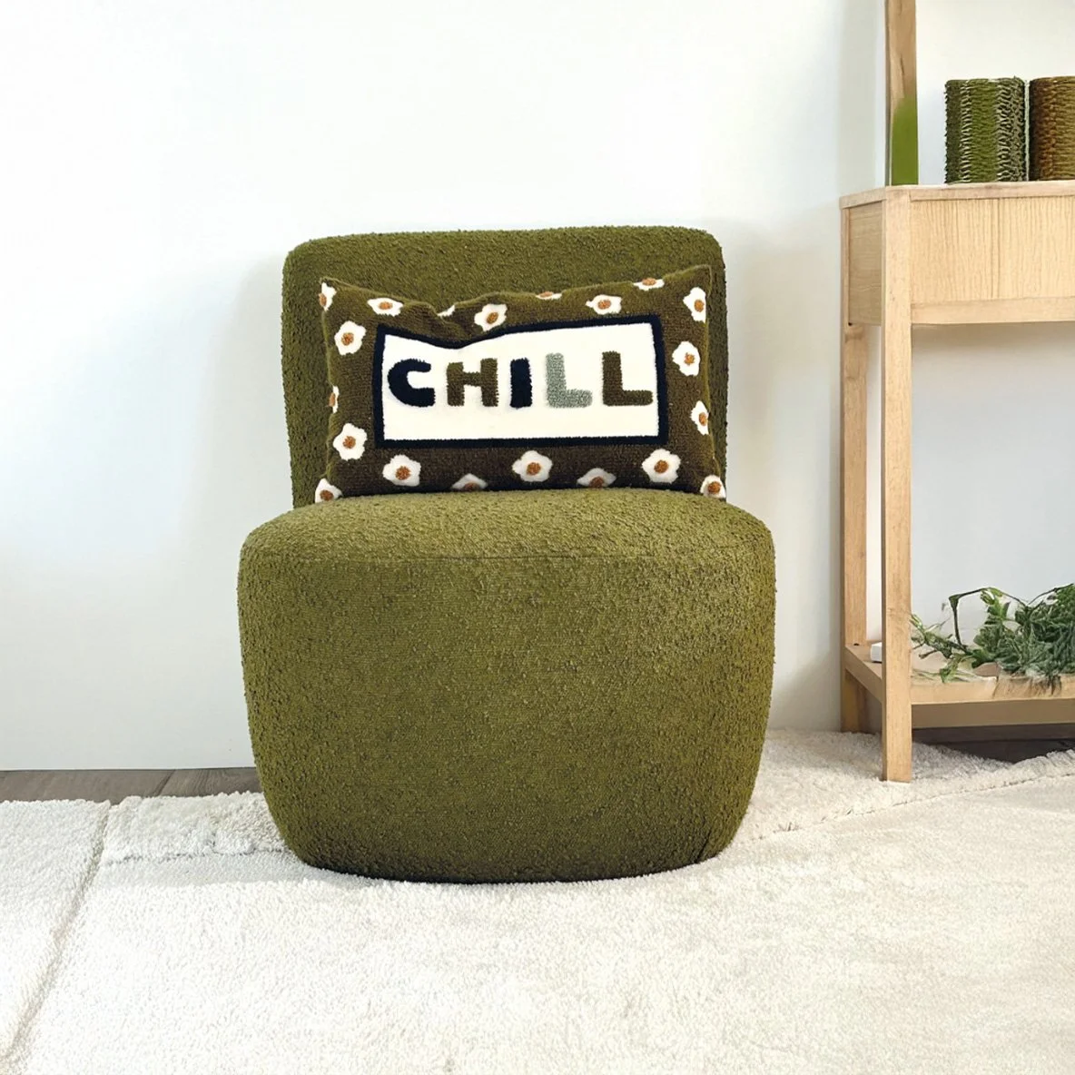 Fauteuil vert bouclé avec un coussin à motif fleurs et texte CHILL, dans un intérieur moderne avec une table en bois et des plantes.
