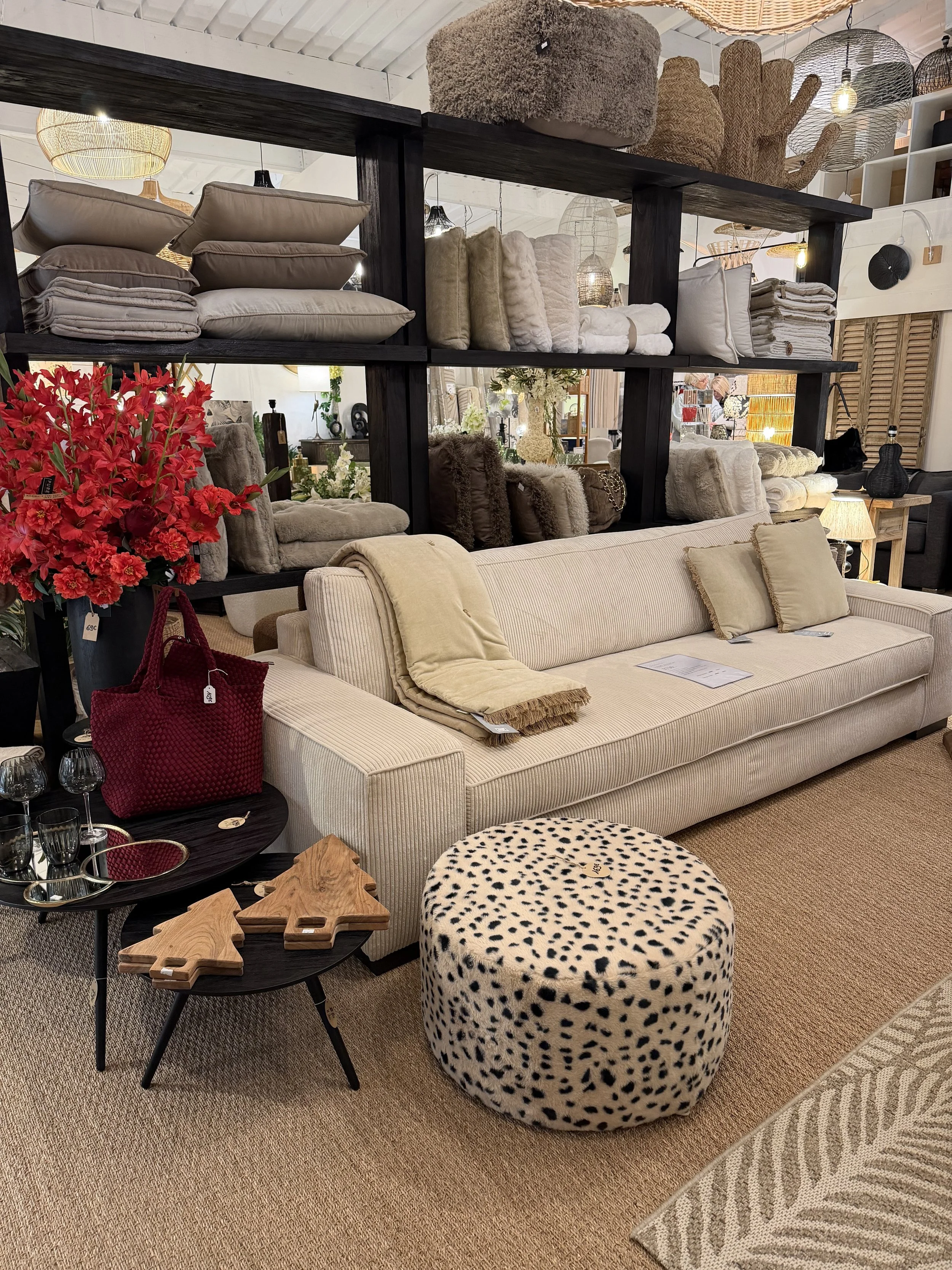 Intérieur d'un magasin de décoration avec un canapé beige, une table basse avec des planches en bois, un pouf à motif animal, des étagères avec des coussins, des couvertures, des lampes, et des accessoires décoratifs.