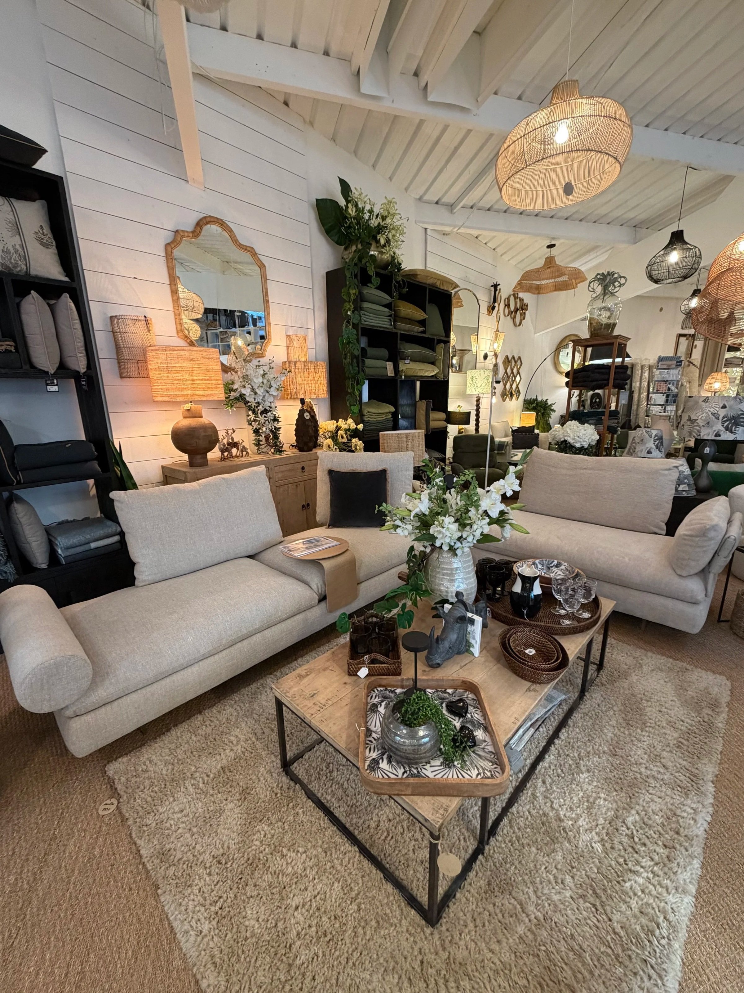 Intérieur d'un magasin de meubles avec deux sofas beige, une table basse en bois avec divers objets décoratifs, un gros vase de fleurs blanches et plusieurs lampes en rotin suspendues au plafond.