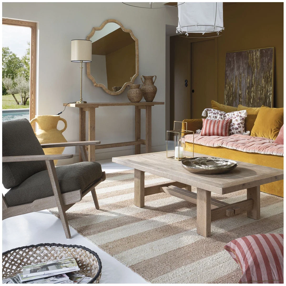 Salon avec canapé jaune, fauteuil gris, table en bois, tapis rayé beige et blanc, décoration murale, lampadaire, miroir, et vue sur jardin par la fenêtre.