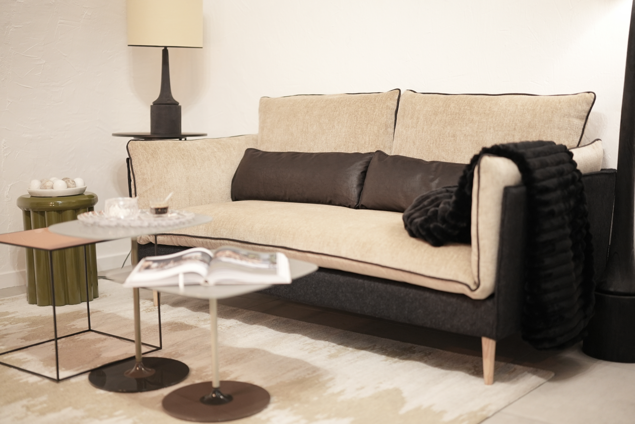 Un canapé beige avec deux coussins marron et une couverture noire dans un salon moderne. Il y a une table en face avec un magazine et deux petites tables à côté, une vert olive et une rose. Une lampe noire est derrière le canapé.