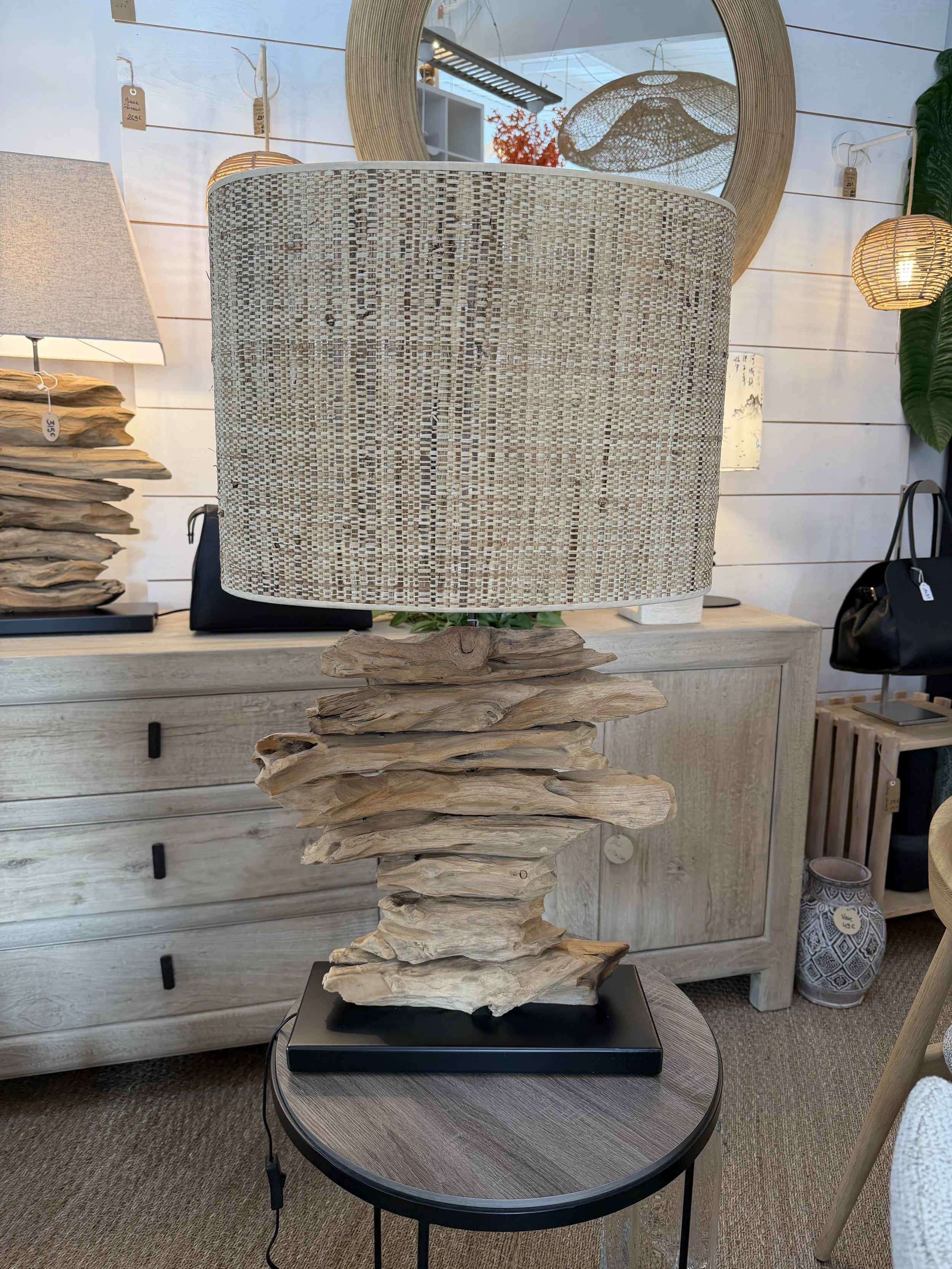 Lampe de table avec base en bois flotté et abat-jour en tissu beige, placée sur une petite table ronde en bois. Décor industriel avec miroir rond, lampes et accessoires en bois et raphia en arrière-plan.