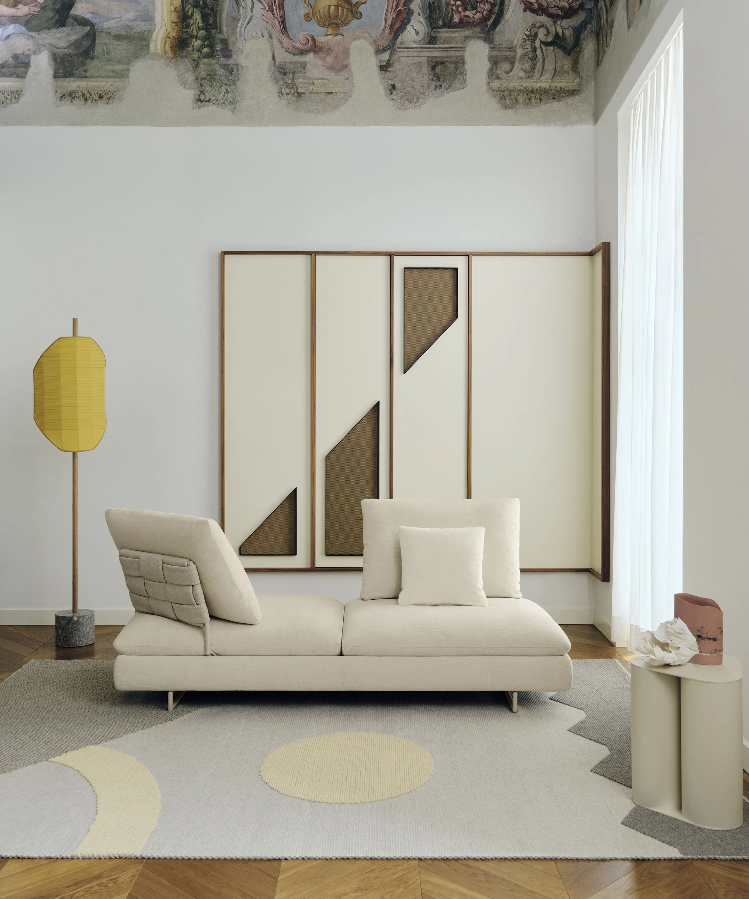 Salon moderne avec un canapé beige, lampadaire jaune, tapis graphique, mur décoratif en bois et tissu, et sculpture abstraite sur une table ronde.