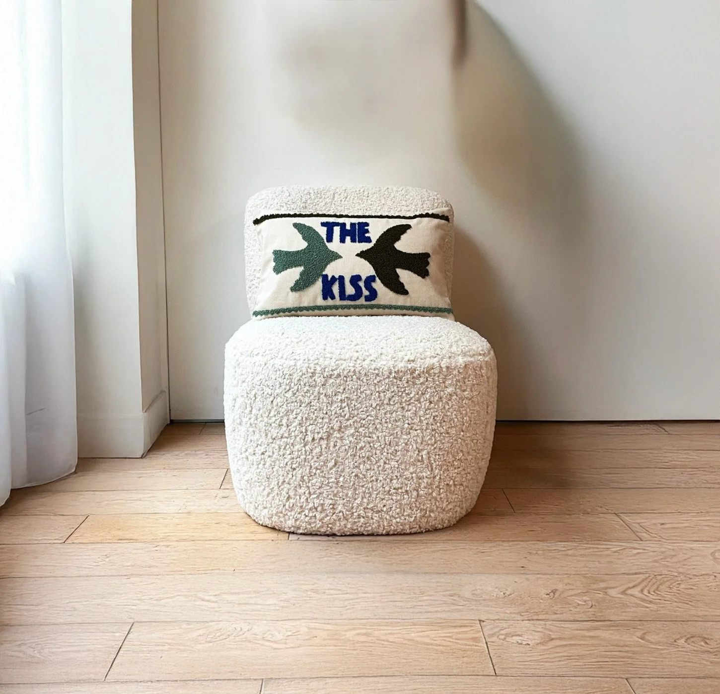 Fauteuil blanc en tissu peluche avec un coussin décoratif brodé avec des oiseaux et l'inscription 'THE KISS', devant un mur blanc et une porte, sur un sol en bois clair.