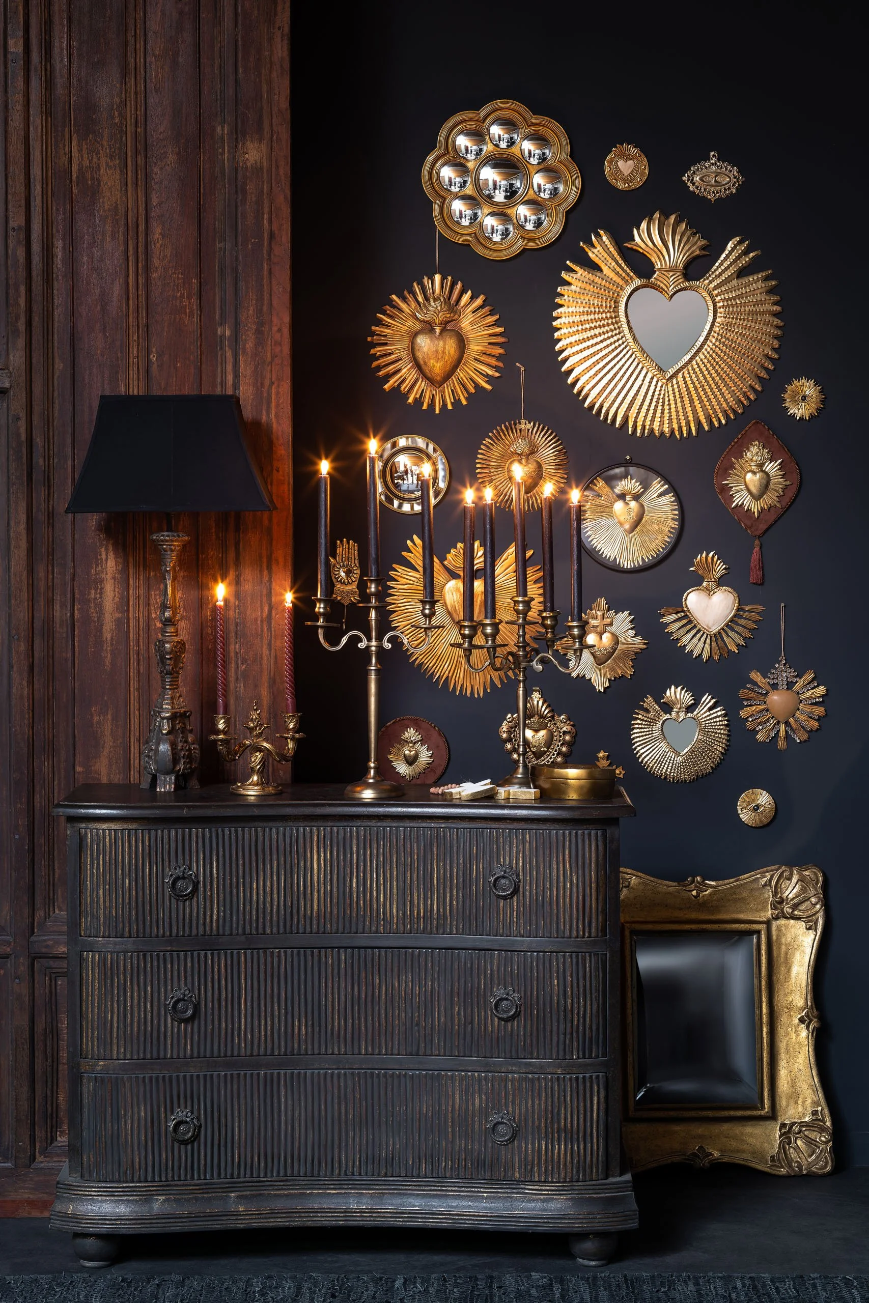 Une commode noire antique avec des tiroirs en détail, décorée de chandeliers en métal avec des bougies allumées et une lampe de bureau noire. Le mur derrière est peint en noir foncé et decoré avec une collection de cadres en forme de soleil, de cœurs et d'autres motifs dorés, créant un style sophistiqué et élégant.