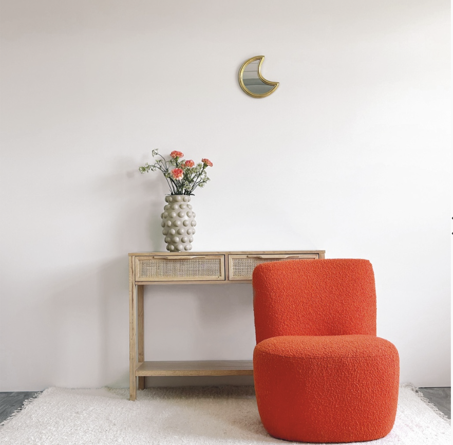 Une pièce minimaliste avec un mur blanc, une console en bois, un vase avec des fleurs, un miroir en forme de lune et un fauteuil orange.