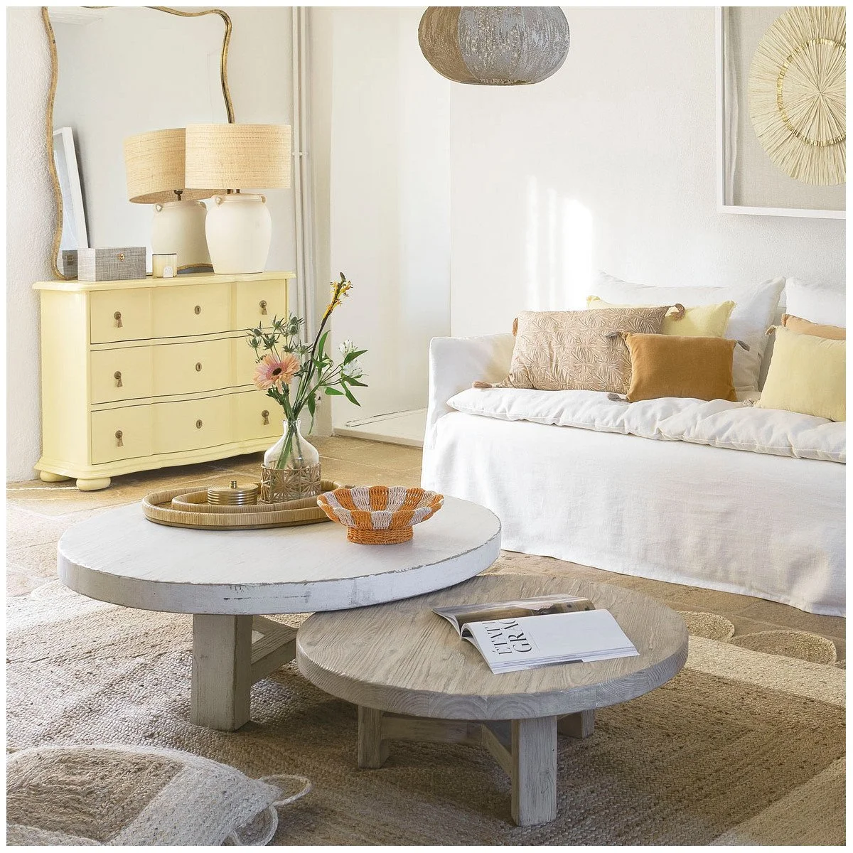 Salon lumineux avec canapé blanc, coussins beiges et jaunes, tables basses en bois, décoration florale, lampes et miroir doré.