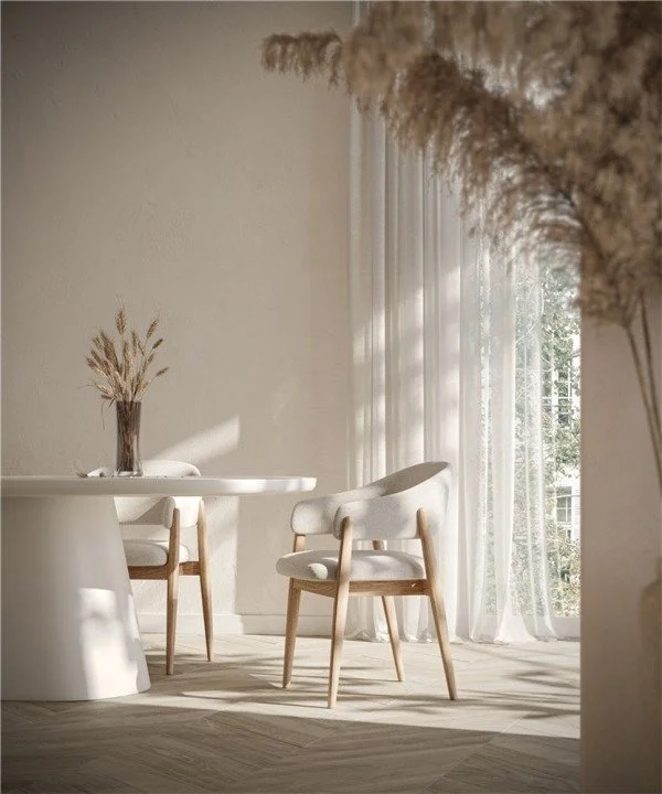 Une salle à manger lumineuse avec une table blanche, deux chaises en bois avec coussins blancs, un vase avec des plantes séchées, des rideaux en lin blanc et une grande fenêtre laissant entrer la lumière