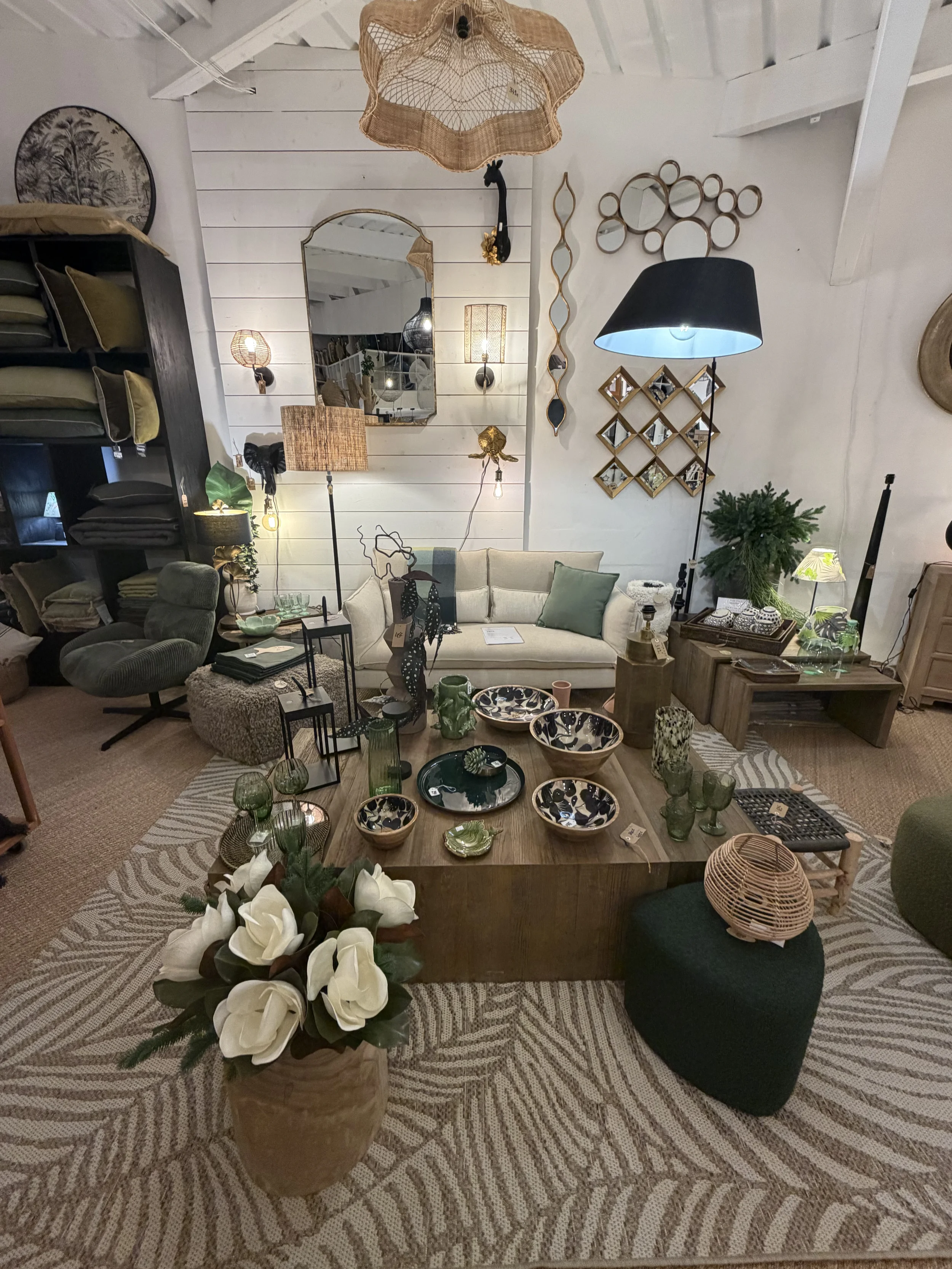 Une salle de séjour décorée avec divers meubles et objets. Il y a un canapé beige, une table basse en bois avec des plats et vaisselle, plusieurs lampes, une étagère à plantes, un miroir, et des décorations murales. Des éléments comme un fauteuil ver