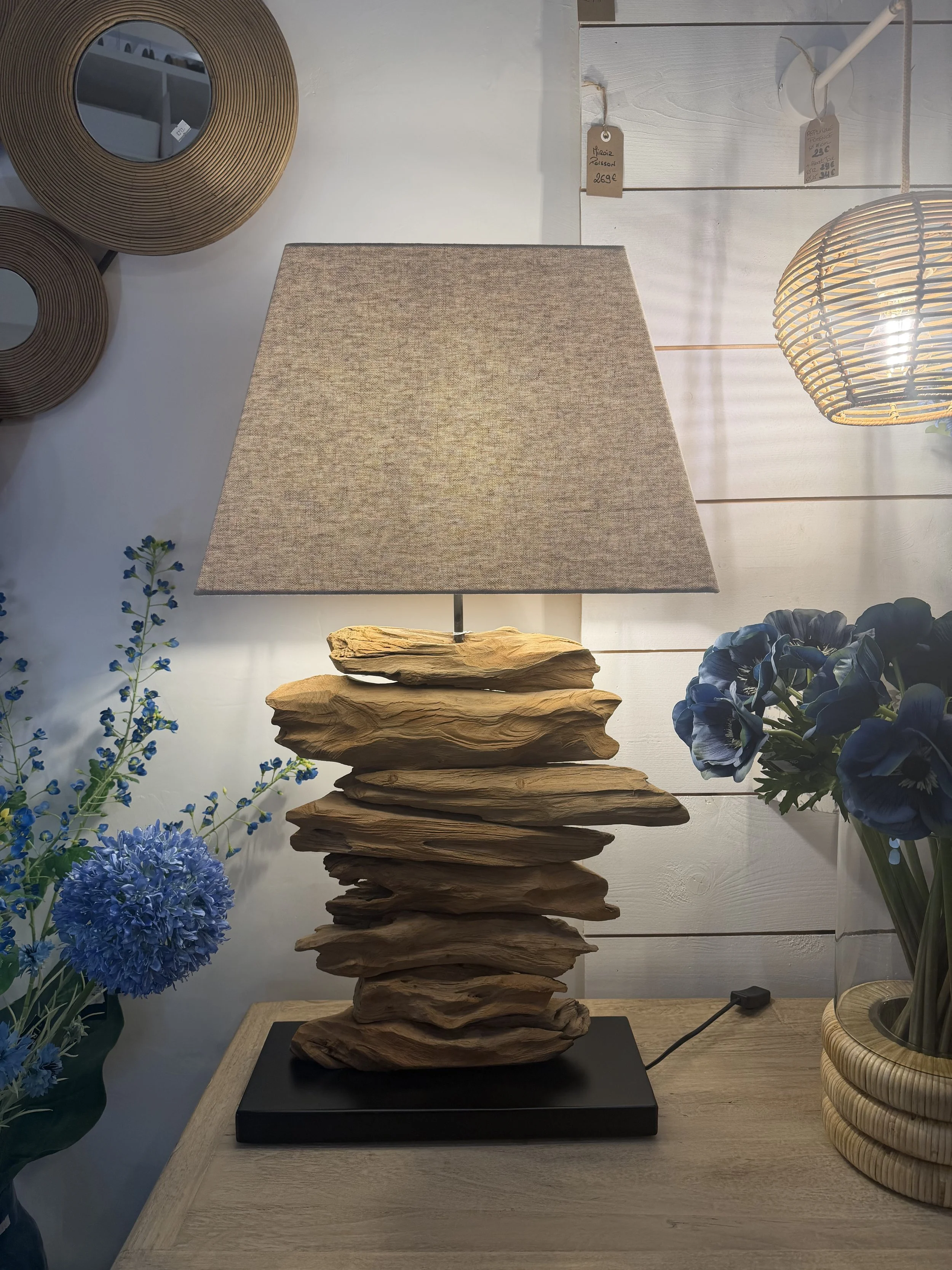 Lampe de table avec un socle en bois naturel, abat-jour en tissu beige, placée sur une table en bois dans un décor intérieur avec des fleurs bleues et des accessoires décoratifs.