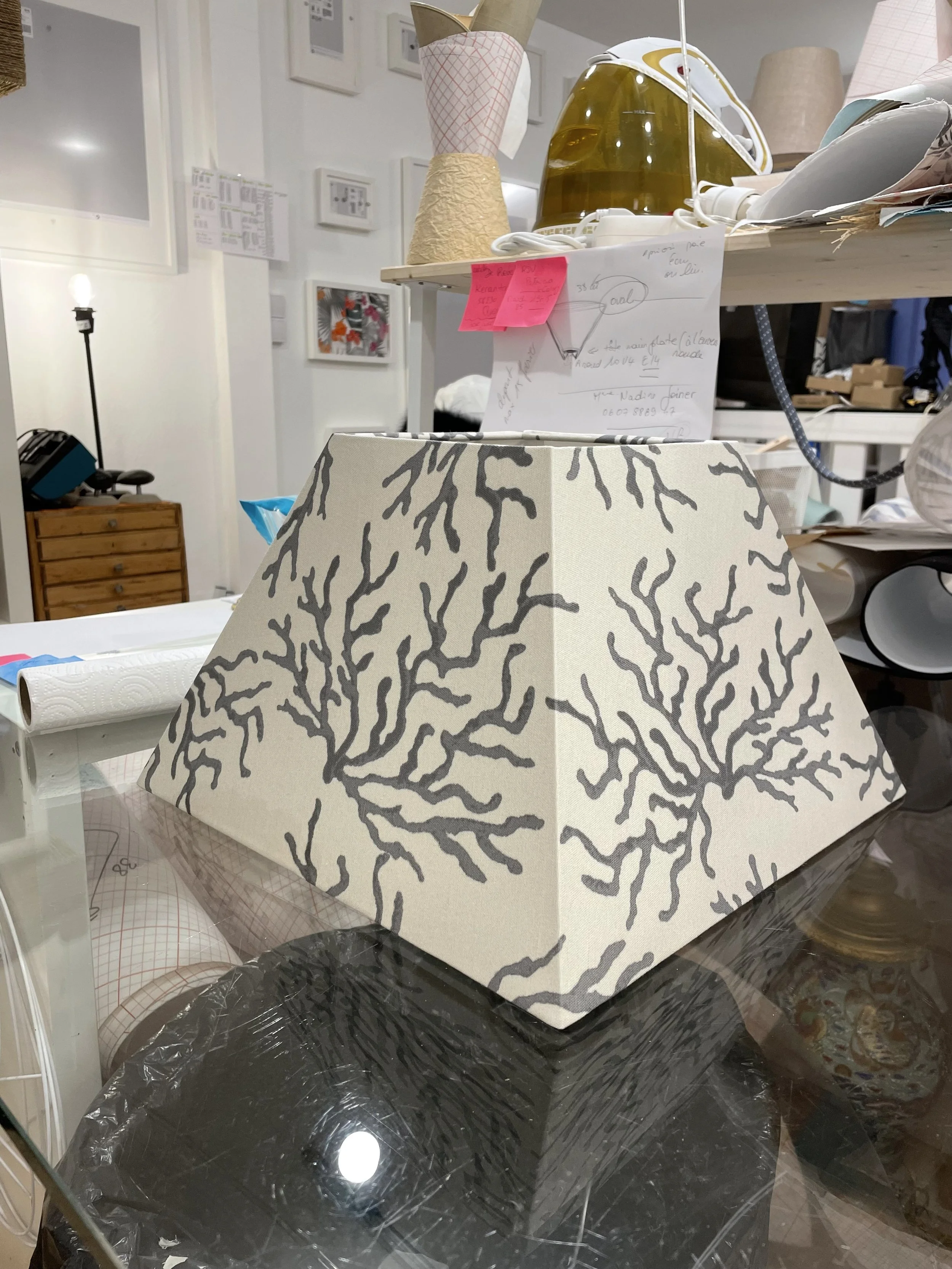 Lampe de table avec abat-jour décoré d'un motif de branches ou d'arbres en gris sur fond blanc, située sur une table en verre.