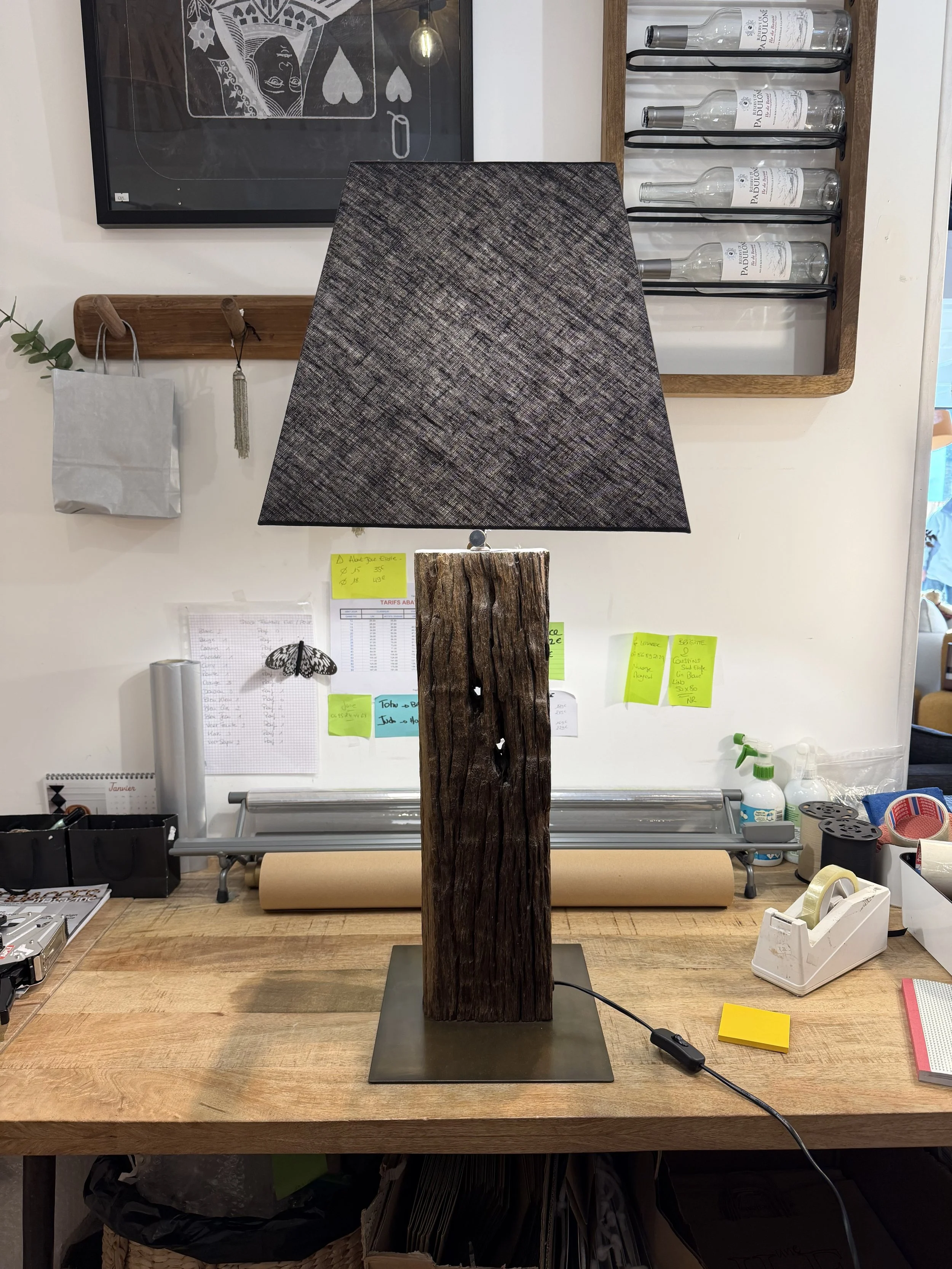 Une lampe de table avec un socle en bois recyclé et un abat-jour en tissu gris, posée sur un bureau en bois.