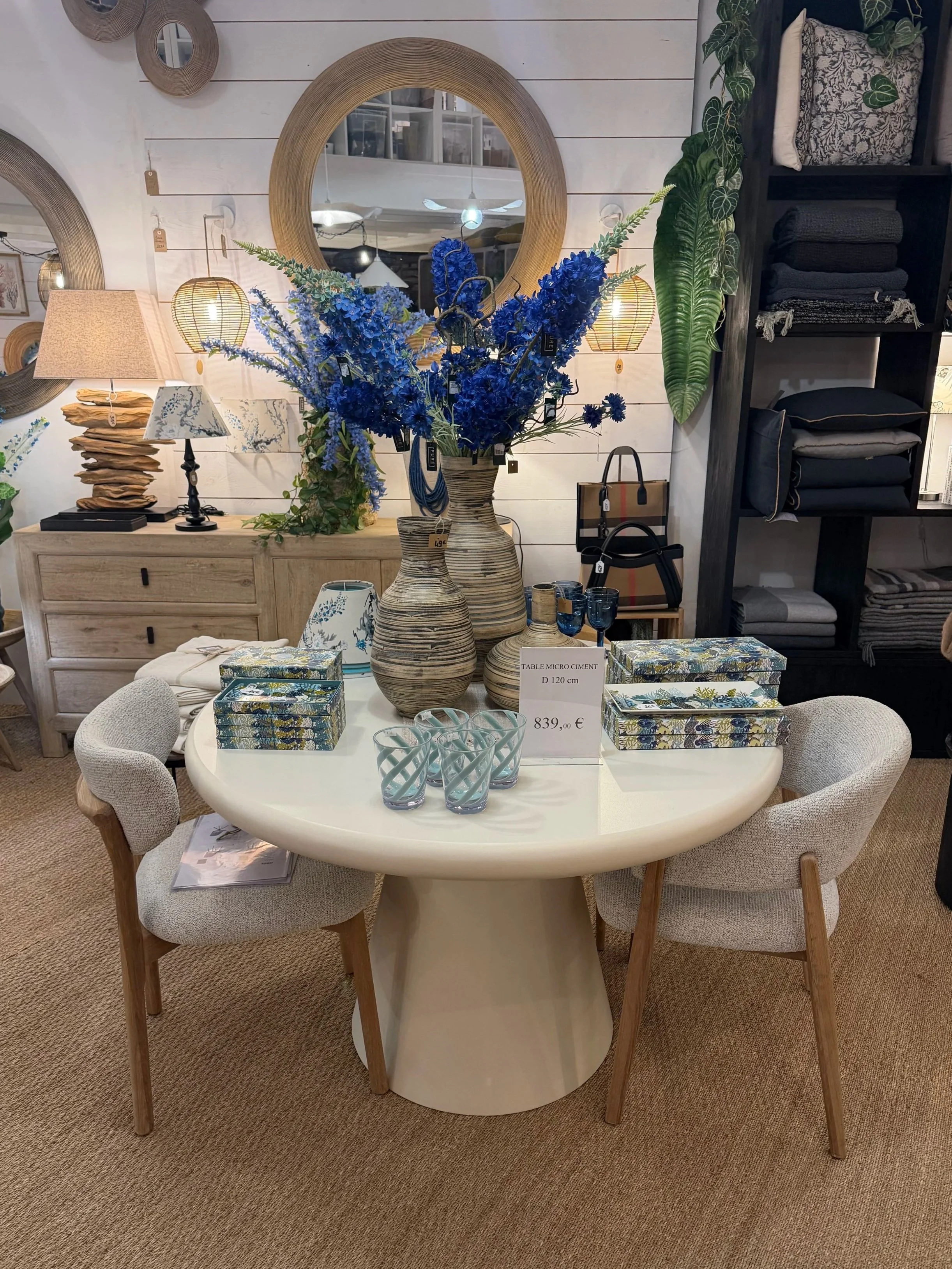 Table ronde blanche décorée avec un vase en céramique contenant des fleurs bleues, entourée de quatre chaises avec des garnitures en tissu beige, dans un magasin de décoration intérieure.