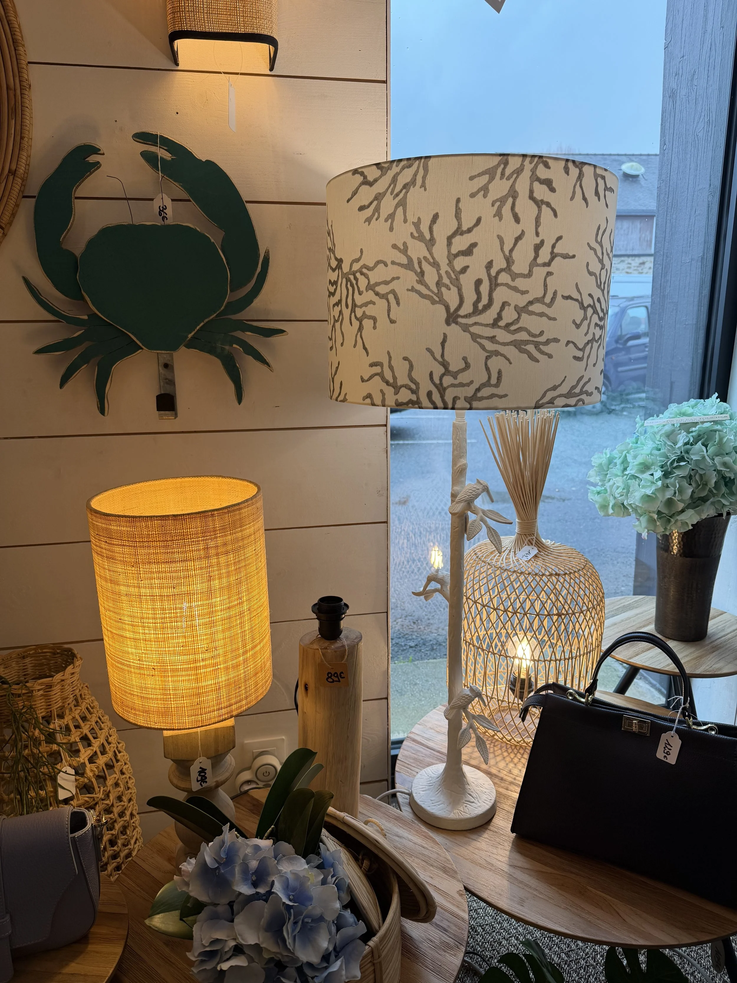Luminaires et décorations d'intérieur dans une boutique, comprenant deux lampes (une avec abat-jour jaune et une avec abat-jour beige à motif de corail), une plante en papier mâché représentant un crabe, une bouteille décorative, un bouquet de fleurs