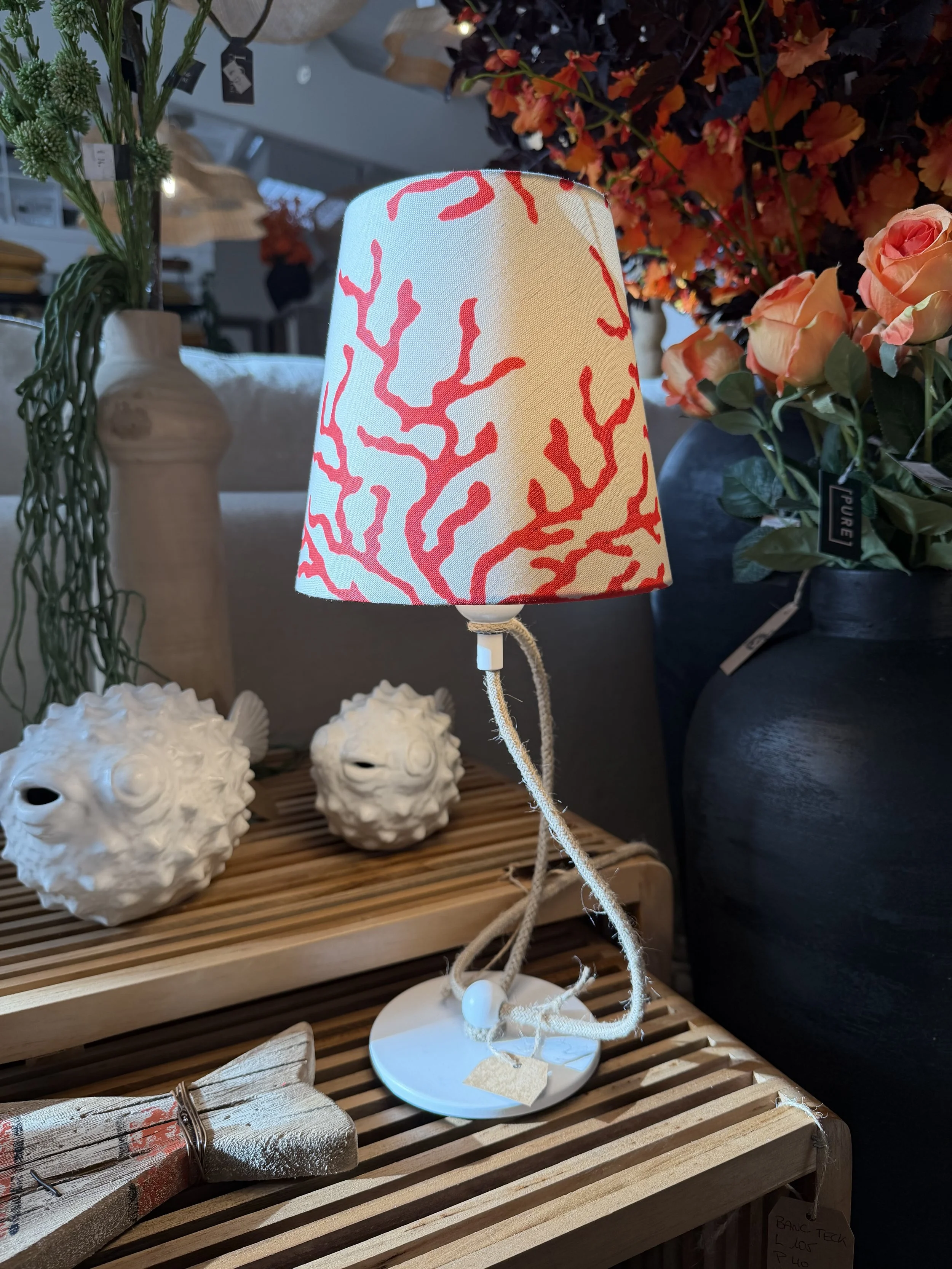 Lampe de table avec abat-jour décoré de motifs de corail rouge, posée sur une table en bois, entourée de déco florale et de céramiques blanches en forme de poissons.