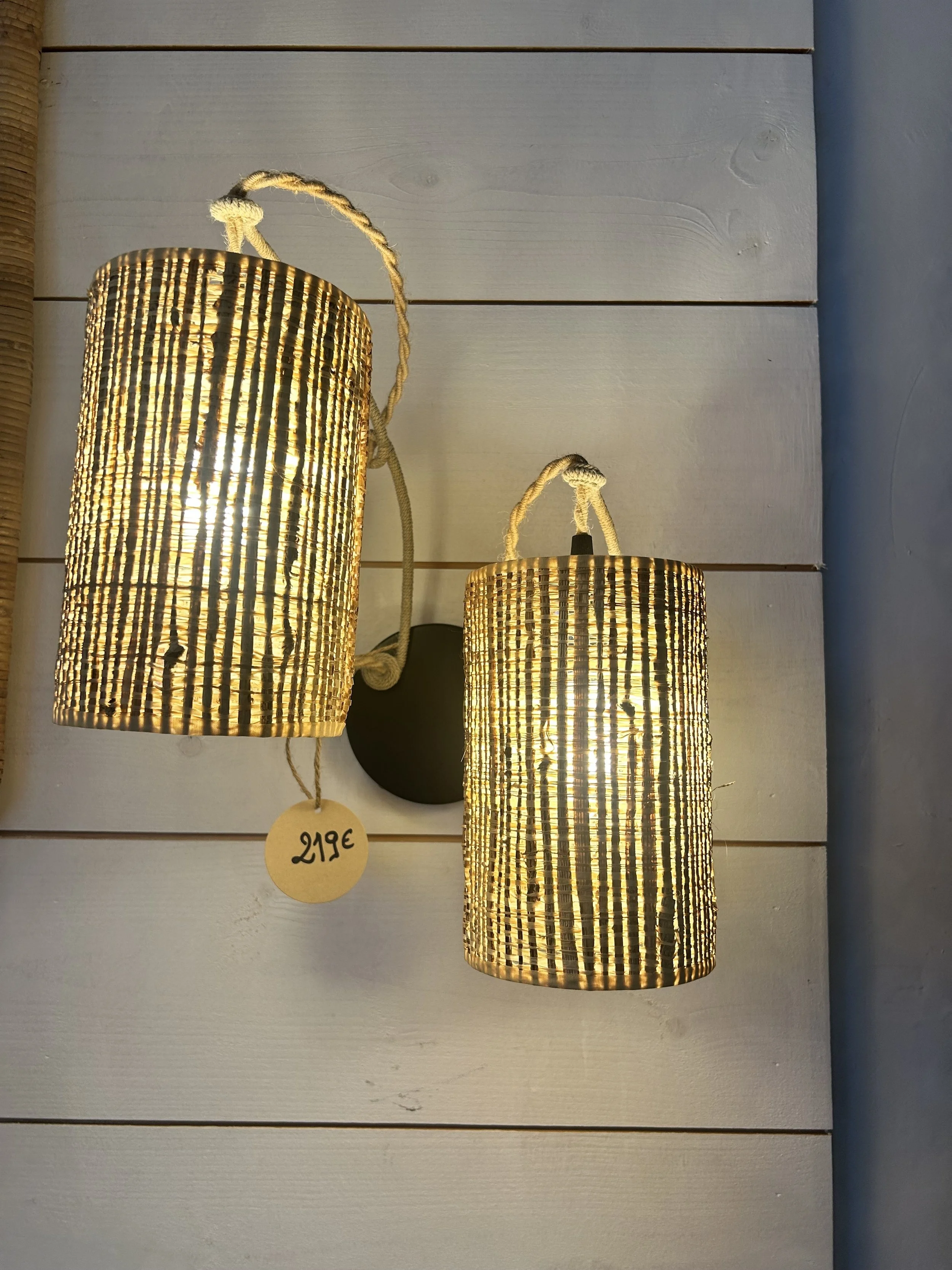 Deux appliques murales avec abat-jour en rotin, illuminant une paroi en bois blanc. Le prix de chaque lampe est indiqué sur un étiquette en papier attachée à la chaîne enroulée autour de l'abat-jour. Le prix indiqué est de 219 €.