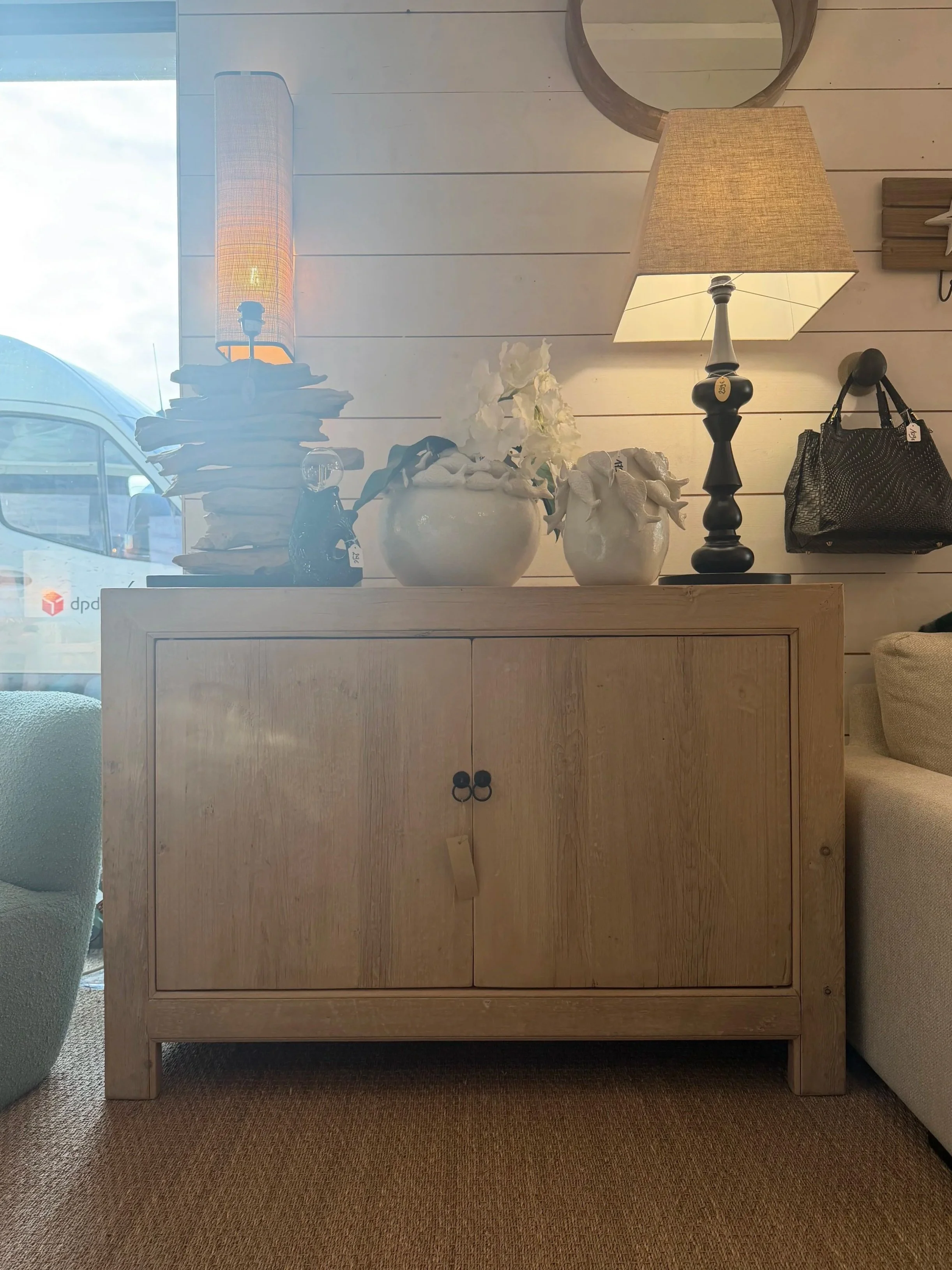 Meuble en bois clair avec deux portes, décoré avec des vases blancs, une lampe à lampe beige et un sac à main noir, dans une salle lumineuse.