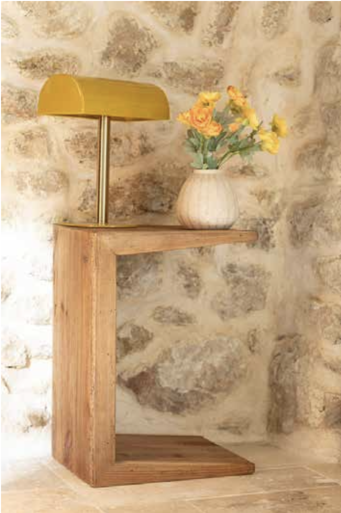 Une lampe de bureau jaune, une vase avec des fleurs jaunes, le tout sur une étagère en bois contre un mur en pierre.