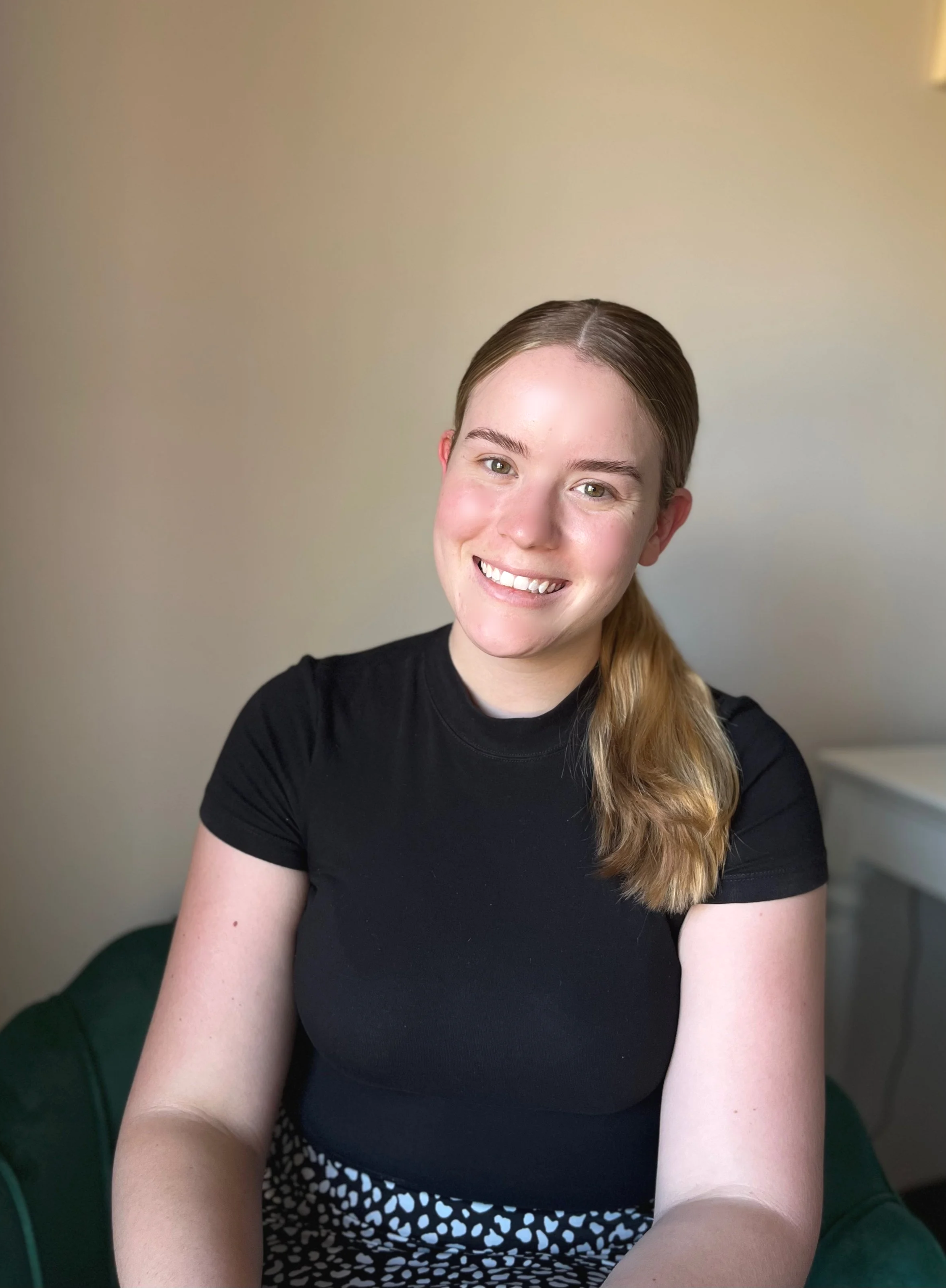 A young woman with long blonde hair tied to the side, wearing a black short-sleeve shirt, smiling while sitting on a green chair in a room with beige walls.