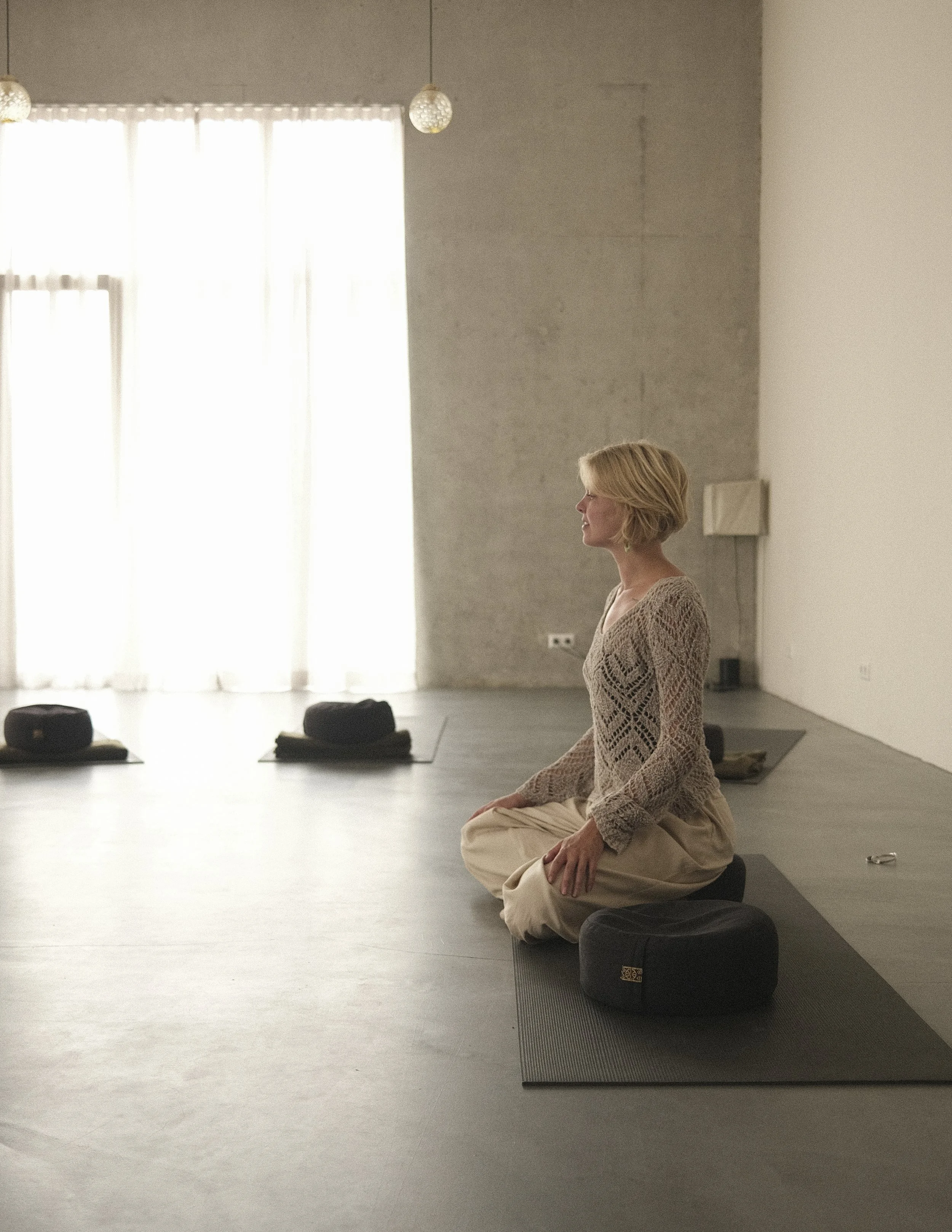 A woman with short blonde hair and glasses sitting on a cushion in a yoga or meditation pose in a bright room with large windows and sheer curtains.