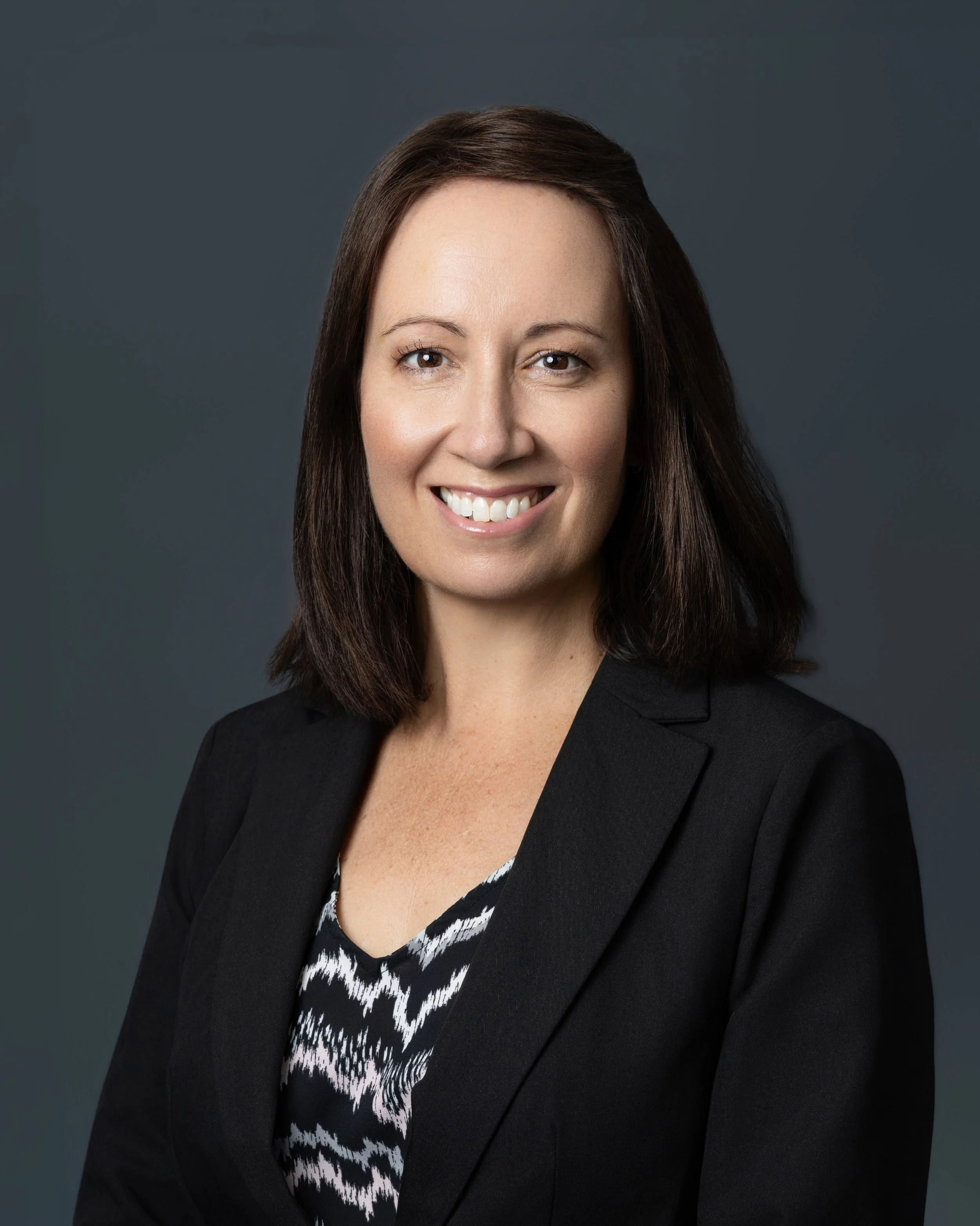 A black and white portrait of a woman with short dark hair, wearing earrings, and smiling at the camera. She is dressed in a patterned blouse and a dark blazer, standing in front of an elevator.