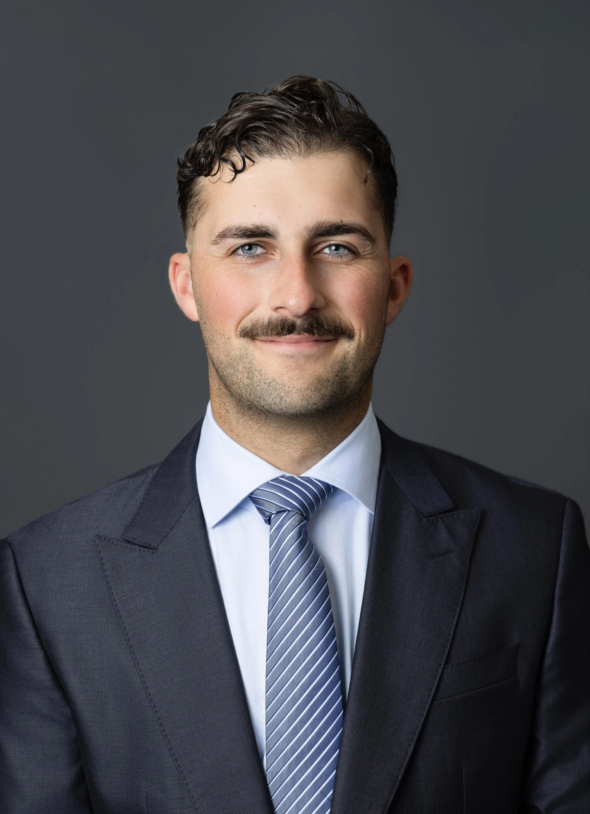 A young man in a dark suit with a white shirt, red tie, and a pocket square, standing outdoors with arms crossed, in front of blurred trees and a wall.