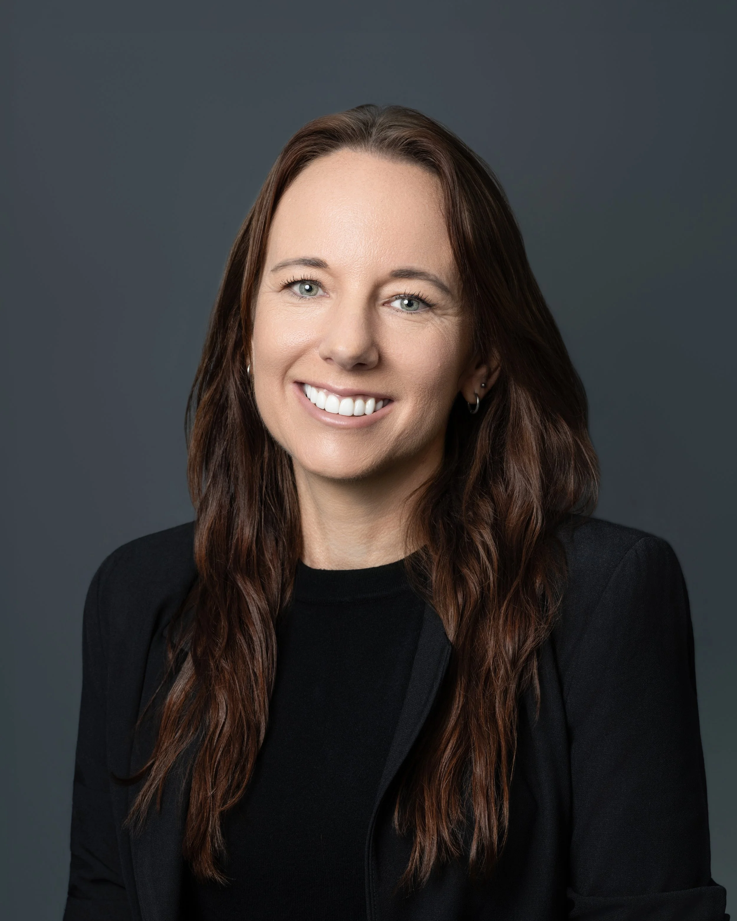 A woman with dark, curly hair styled in a messy bun, smiling and wearing a white t-shirt against a plain white background.