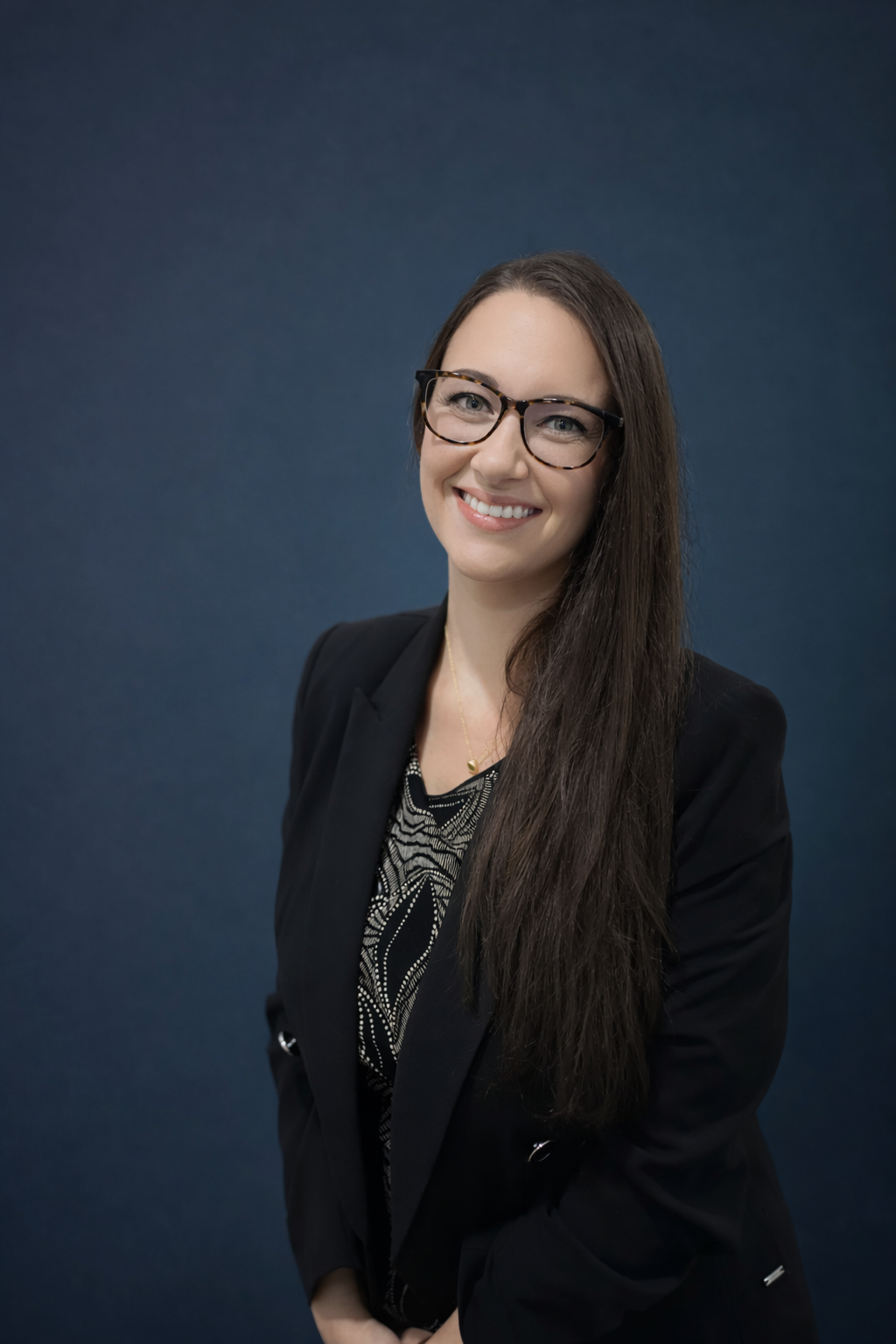 A woman with long dark hair, glasses, and a bright smile, wearing a dark blazer over a patterned top, standing against a plain wall.