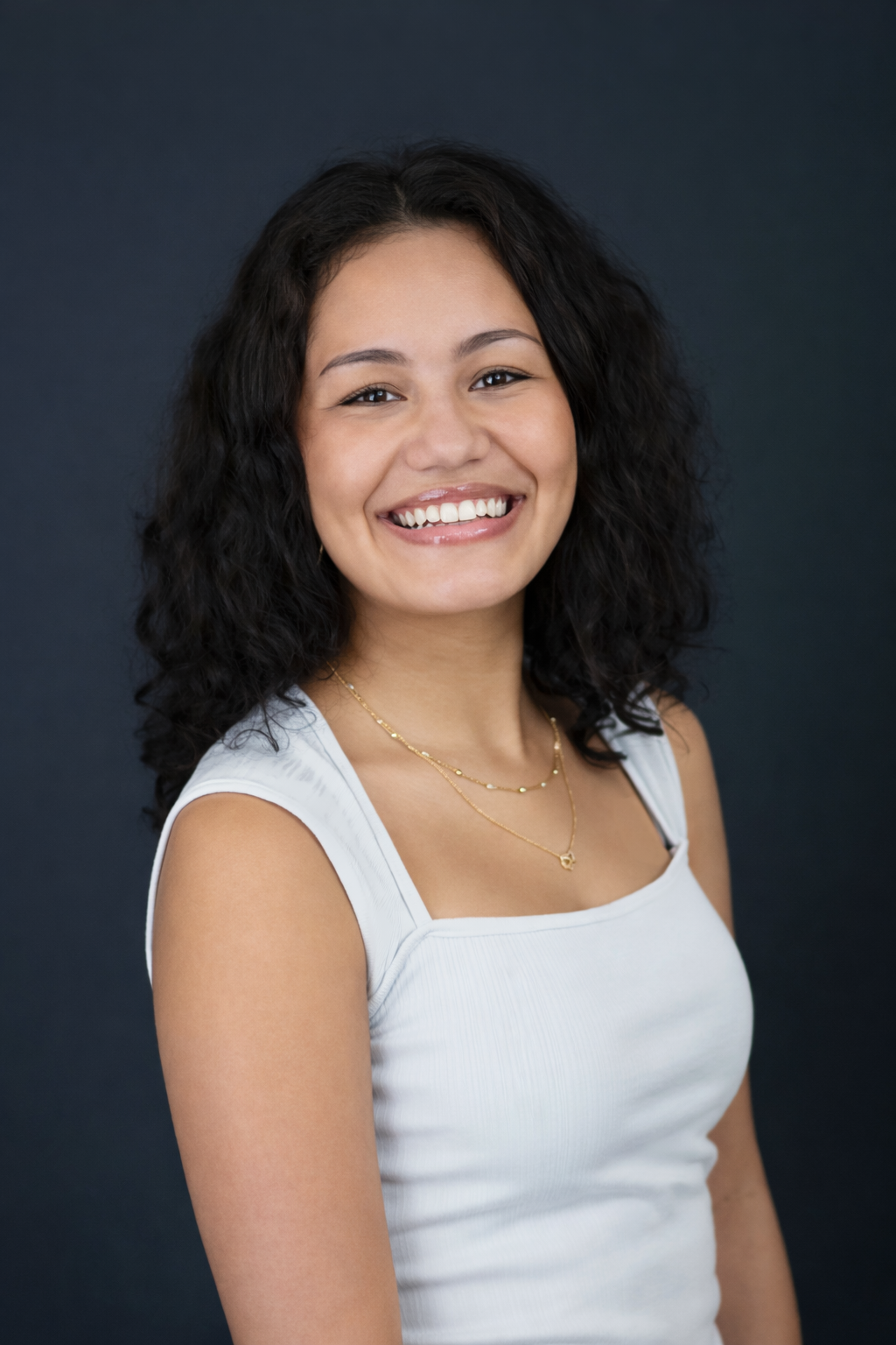 Black and white portrait of a woman with long, wavy dark hair, smiling, wearing a dark blazer over a light top, standing against a plain wall.
