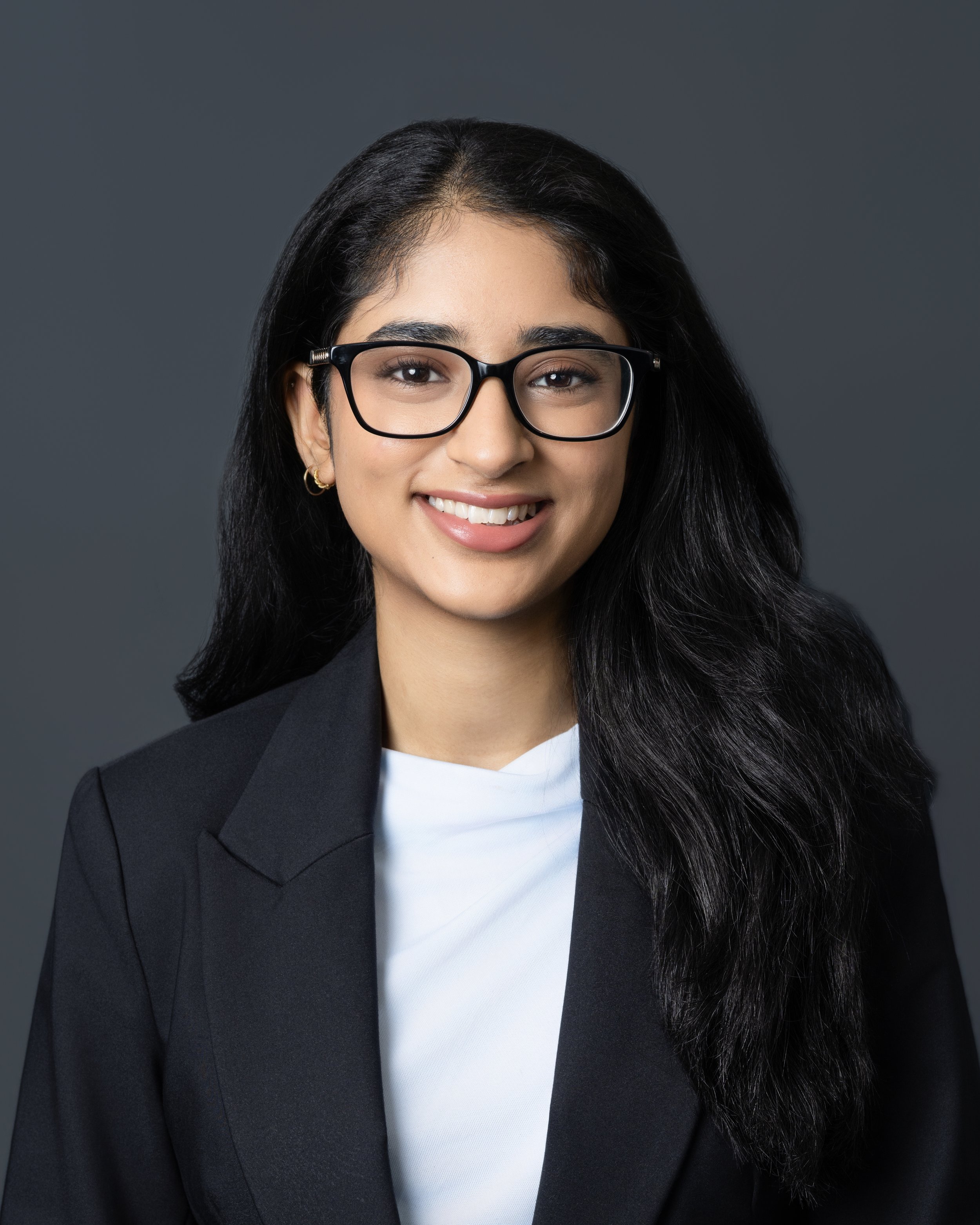 A young woman with long dark hair, glasses, and earrings, smiling, wearing a blazer and a top, standing against a plain wall.