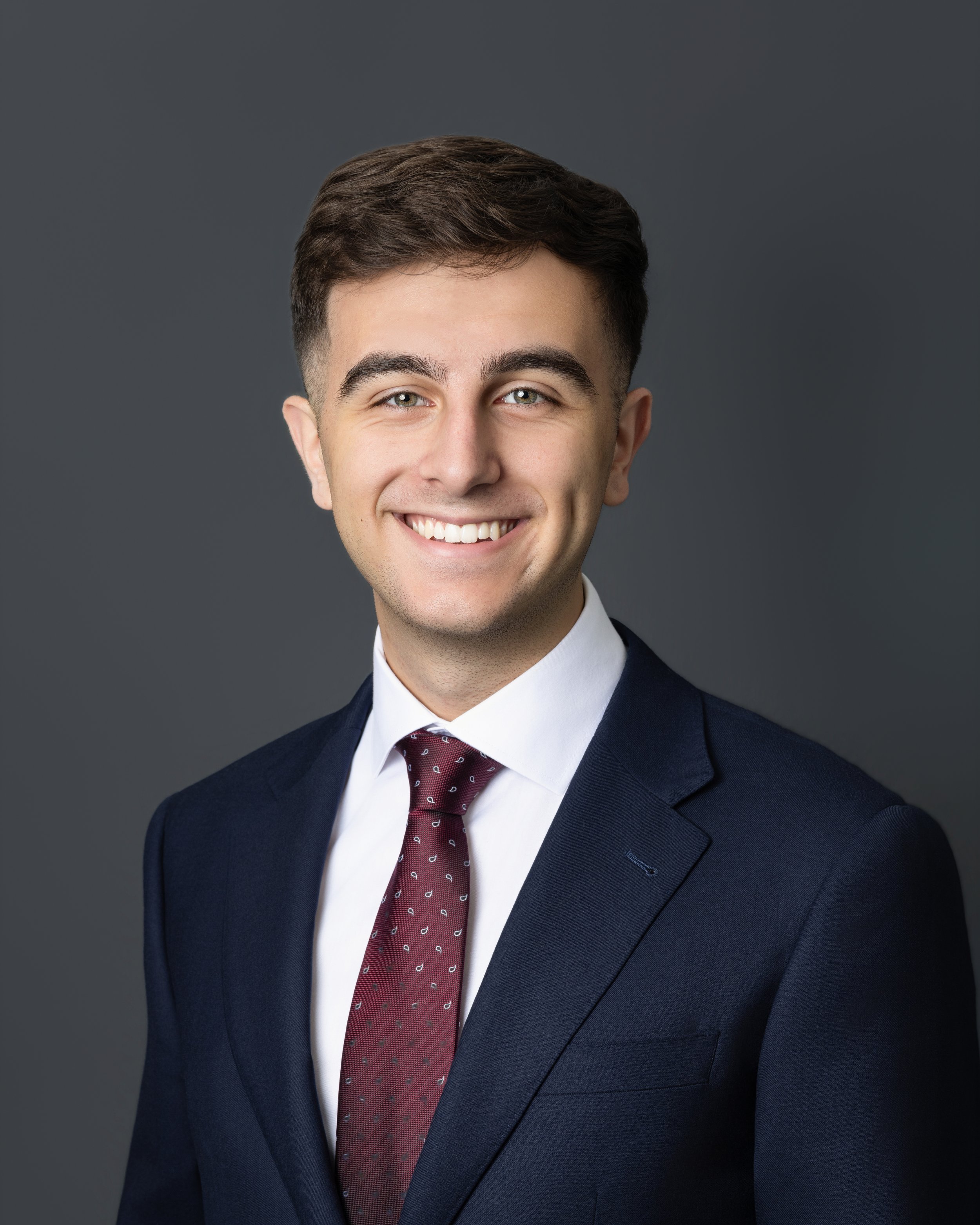 A young man with short hair and a suit, smiling. He is standing in front of a dark wooden door and a plain wall.